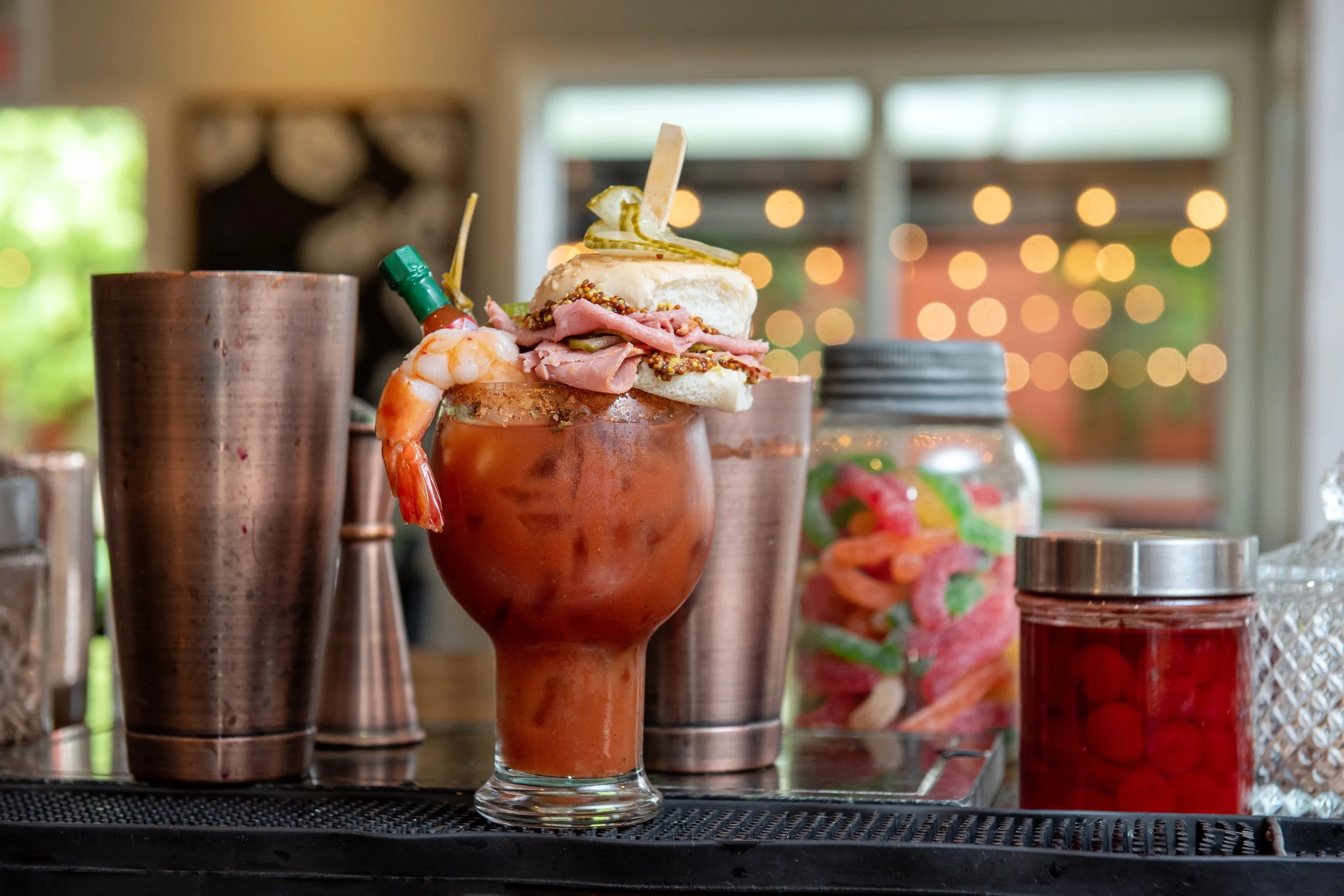 A colorful cocktail garnished with pickles, shrimp, and a slice of lemon, with a small bottle of hot sauce attached to the glass, on a bar counter with various bar tools and bottles in background.