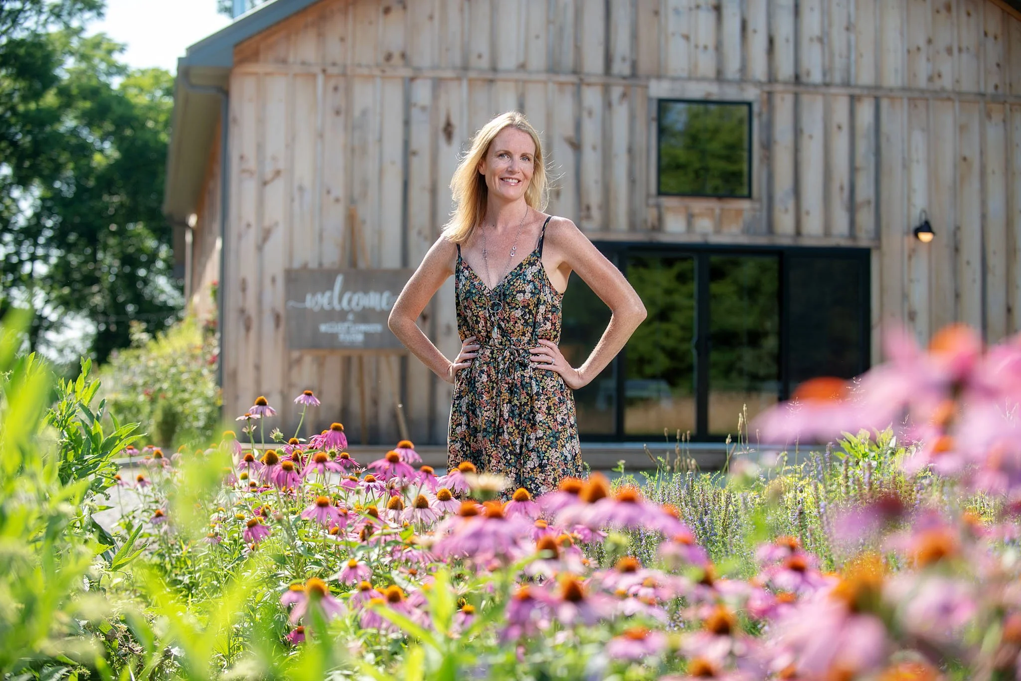 A woman with blonde hair wearing a summer dress standing in a garden with colorful flowers in front of a wooden building with large windows.