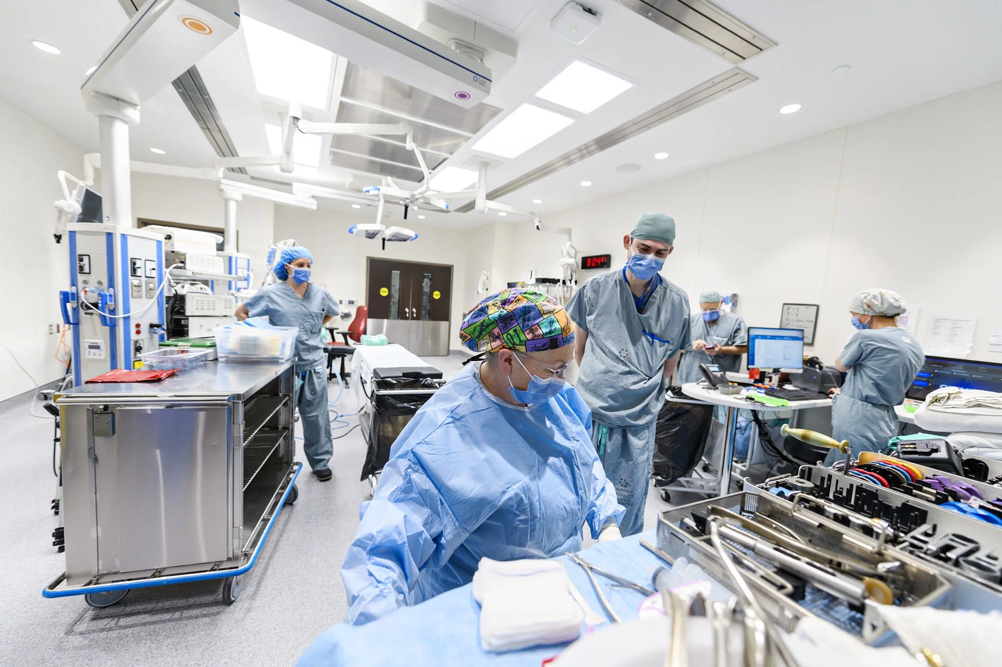 Medical team in an operating room wearing scrubs, masks, and caps, working with surgical instruments and medical equipment.