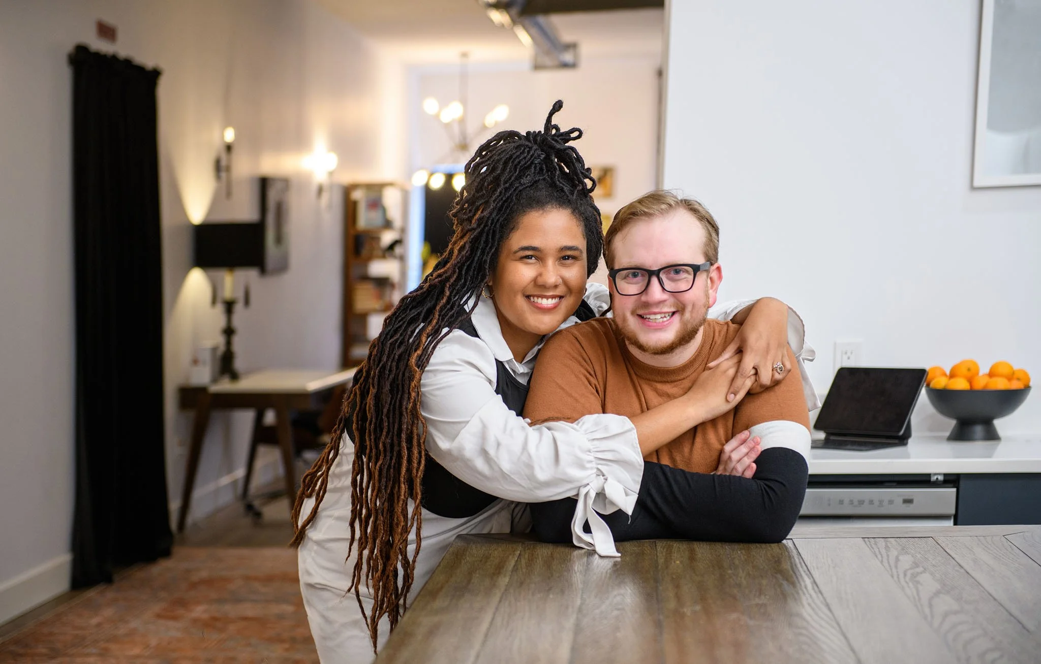 A happy woman with long dreadlocks hugging a smiling man with glasses at a kitchen island with a bowl of oranges and a tablet, in a modern home interior.