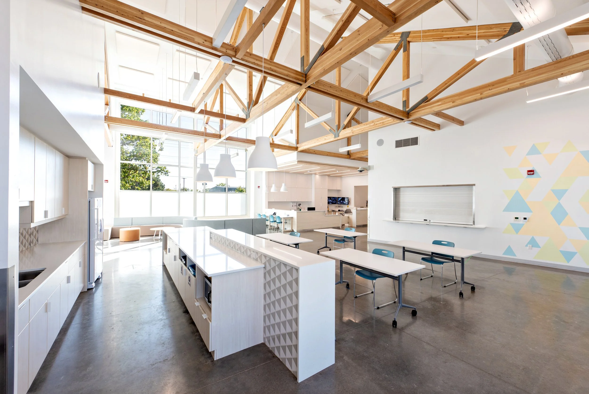 A spacious modern interior with high ceilings, large windows, wooden beams, white walls, pendant lighting, and tables with chairs, likely a communal or meeting area.