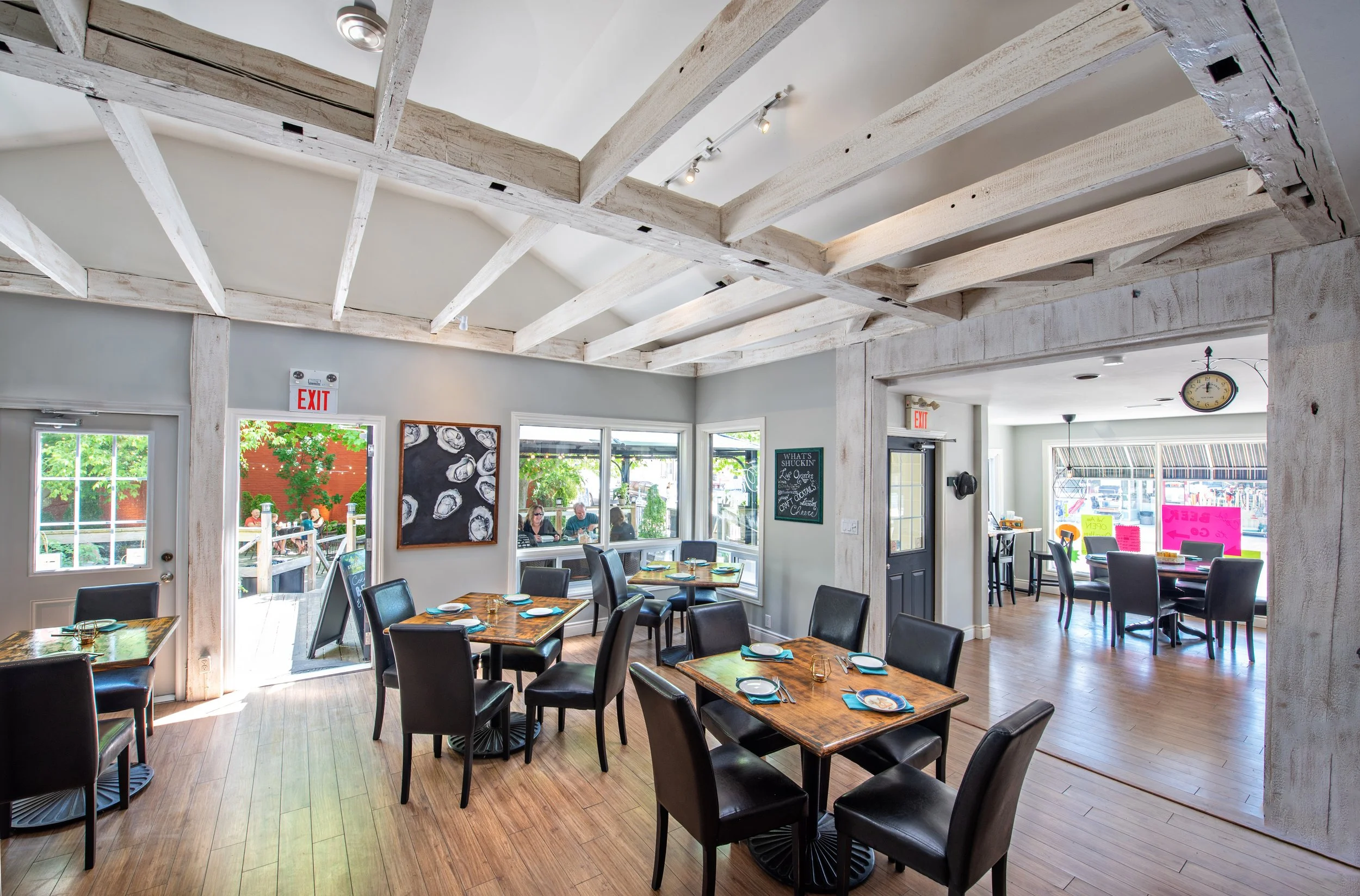 Interior of a restaurant with wooden beams on the ceiling, dining tables with black chairs, large windows allowing natural light, and people seated outside.
