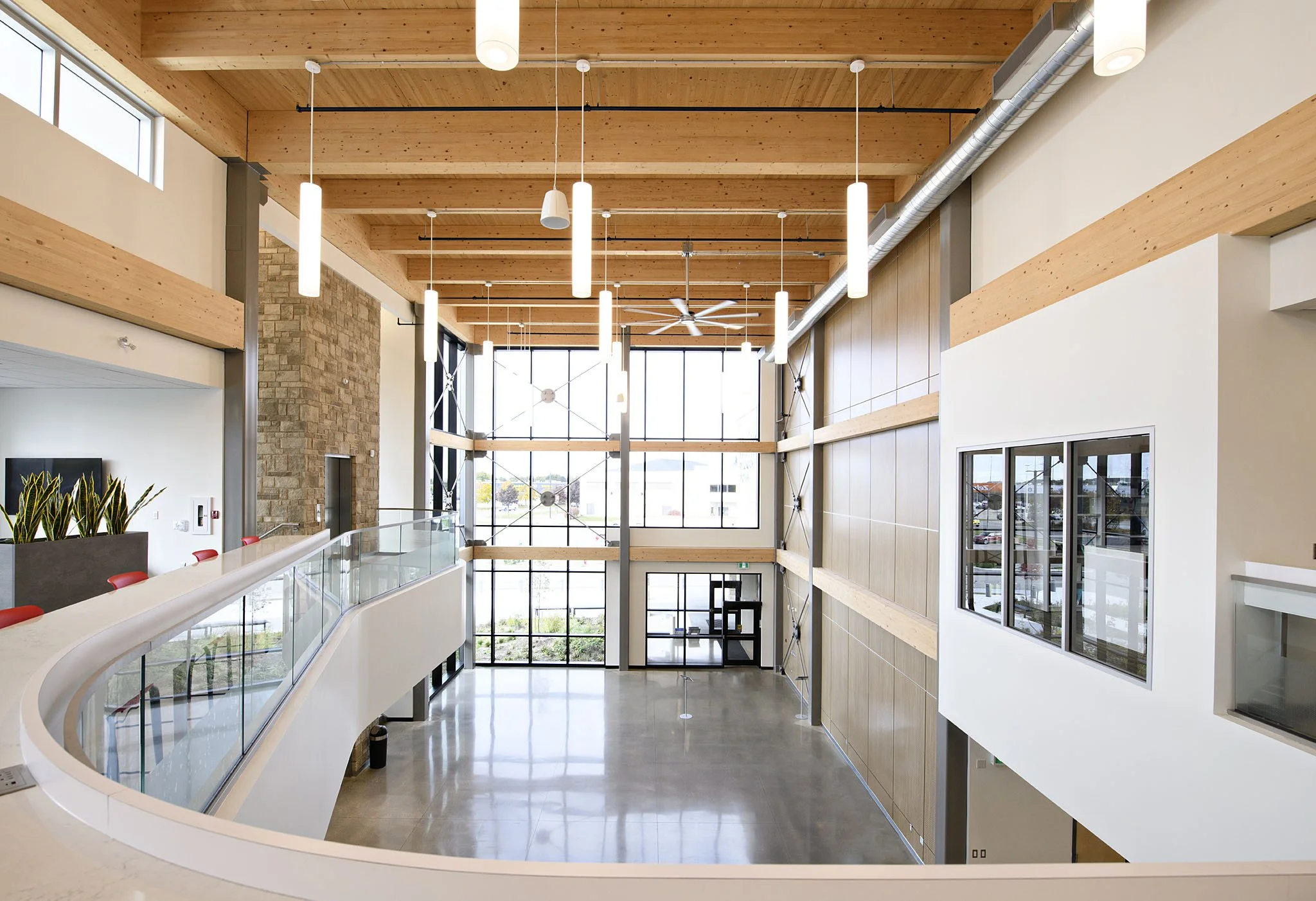 Interior view of a modern building lobby with high ceiling, wooden beams, hanging ceiling lights, large windows, a stone wall on the left, and a glass railing on the second floor.