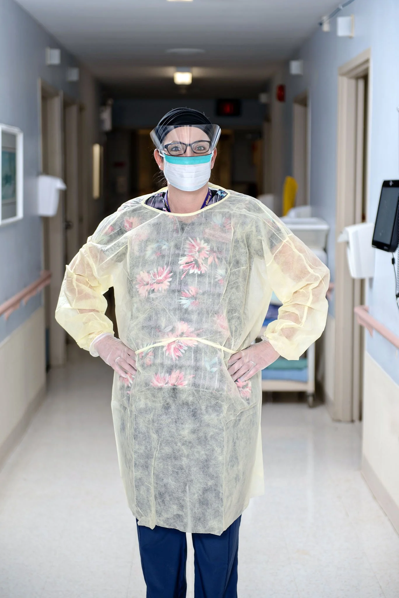 Female healthcare worker standing in hospital corridor wearing medical mask, face shield, protective gown, gloves, and scrubs.