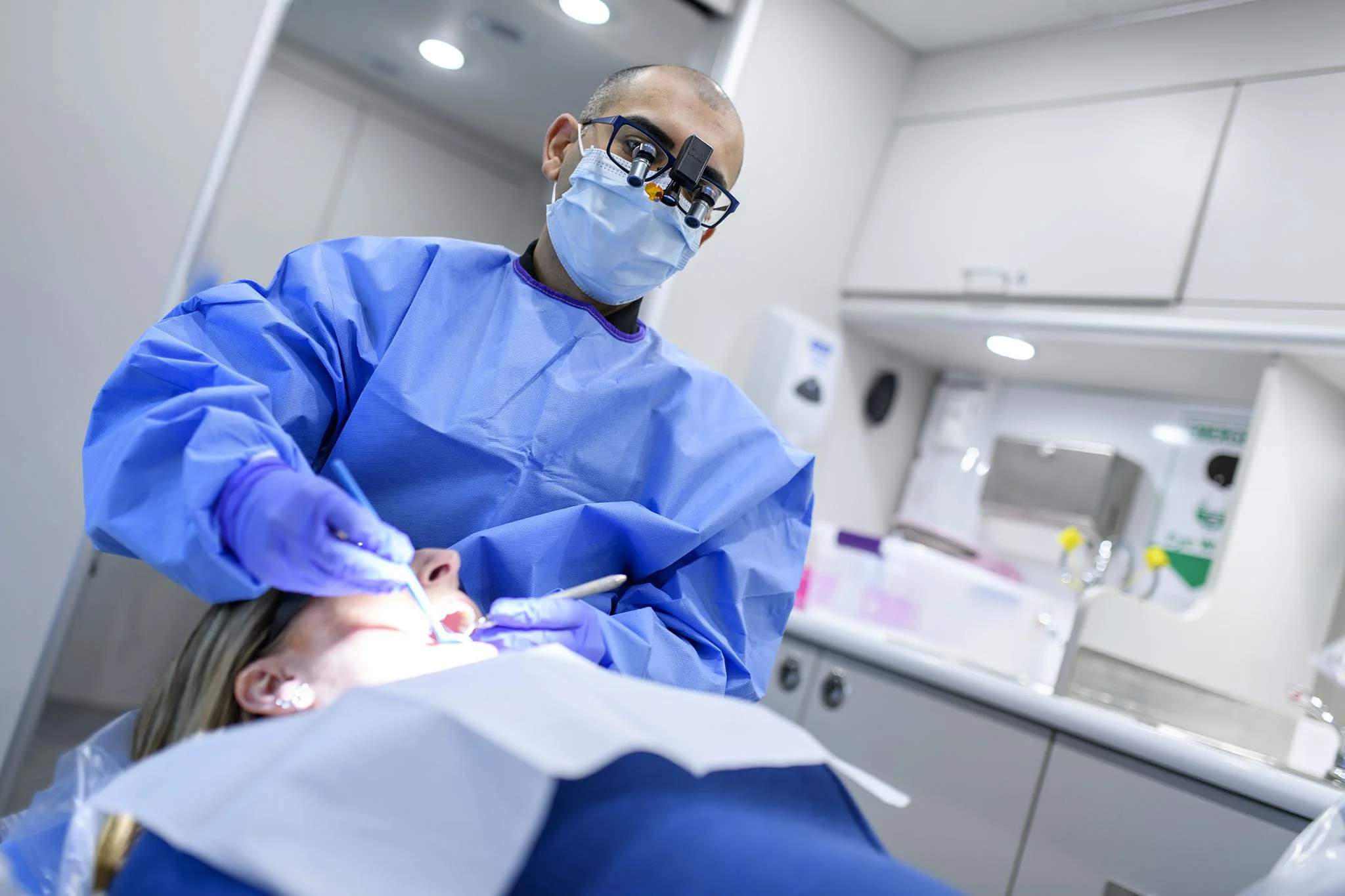 A healthcare professional wearing surgical scrubs, a mask, and magnifying loupes performs a procedure on a patient in a clinical setting, with medical equipment visible in the background. Commercial photography. 