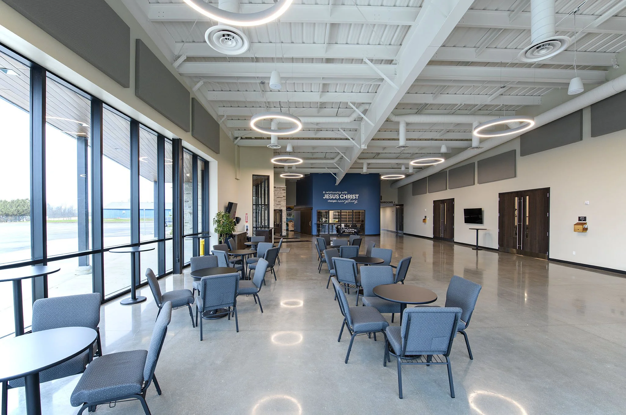 Empty lounge area inside a church or community center with chairs, tables, large windows, and a sign that says "A relationship with Jesus Christ changes everything."