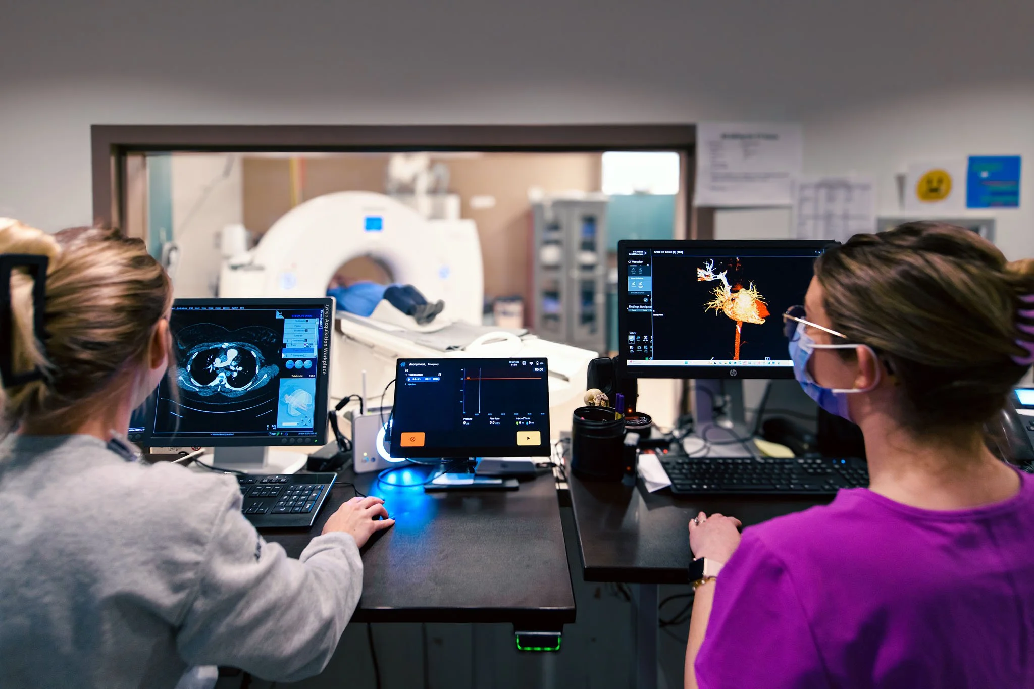 Two medical professionals in masks working at computer monitors in a hospital control room, with a patient undergoing a scan in the background.