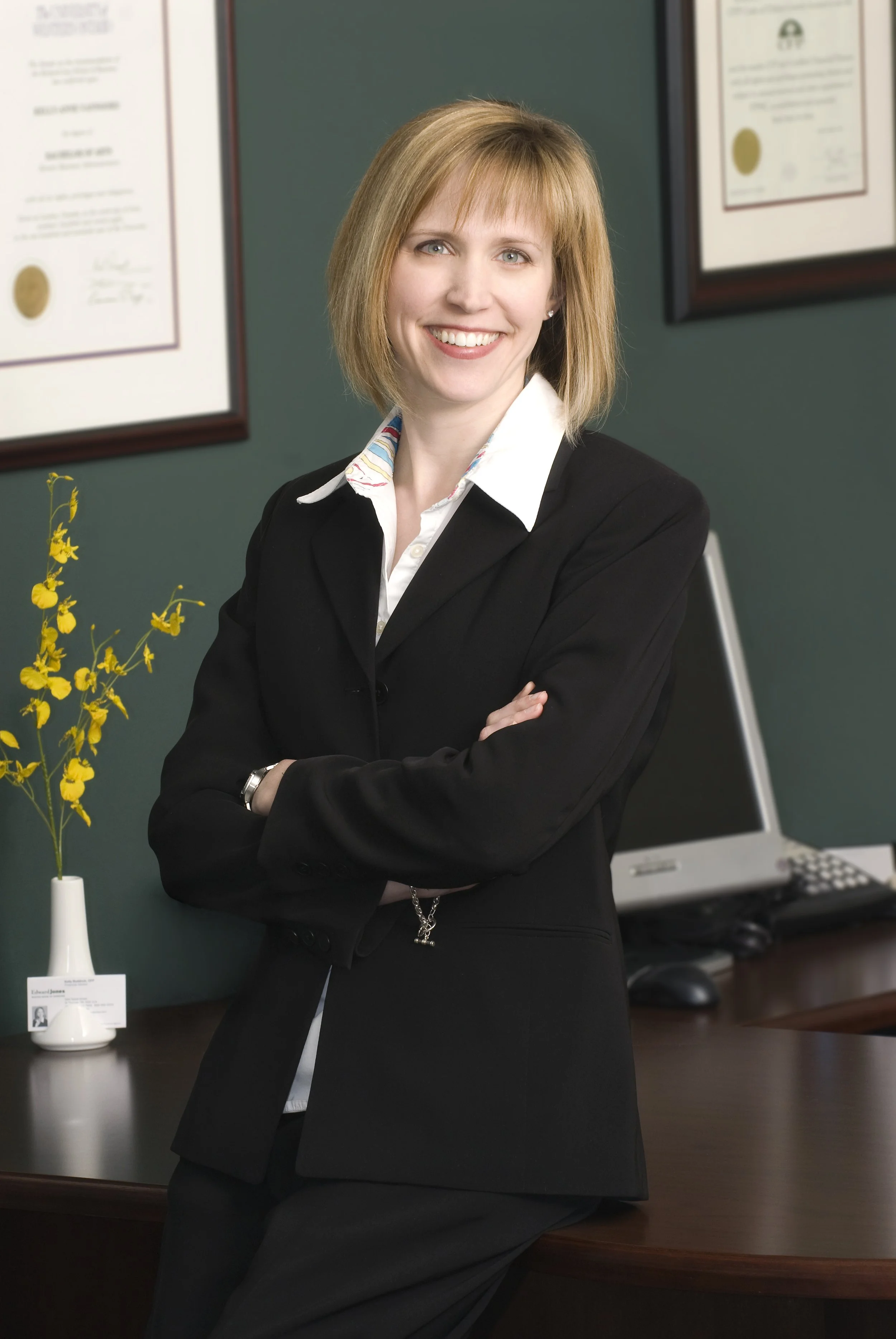 Executive portrait photography. A professional woman with shoulder-length blonde hair, wearing a black blazer and white shirt, smiling with crossed arms in an office setting with framed certificates on the wall and a computer monitor.