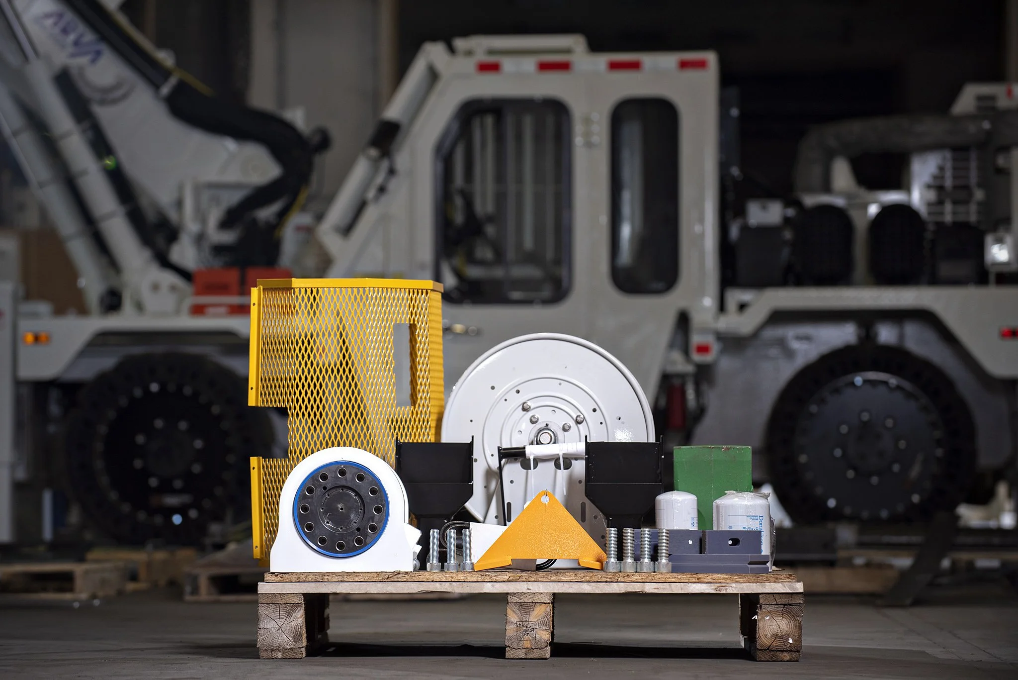 Various mechanical and electronic components, including a yellow mesh cover, white circular parts, and other metal and plastic pieces, placed on a wooden pallet in an industrial setting, with large machinery in the background. Product photography