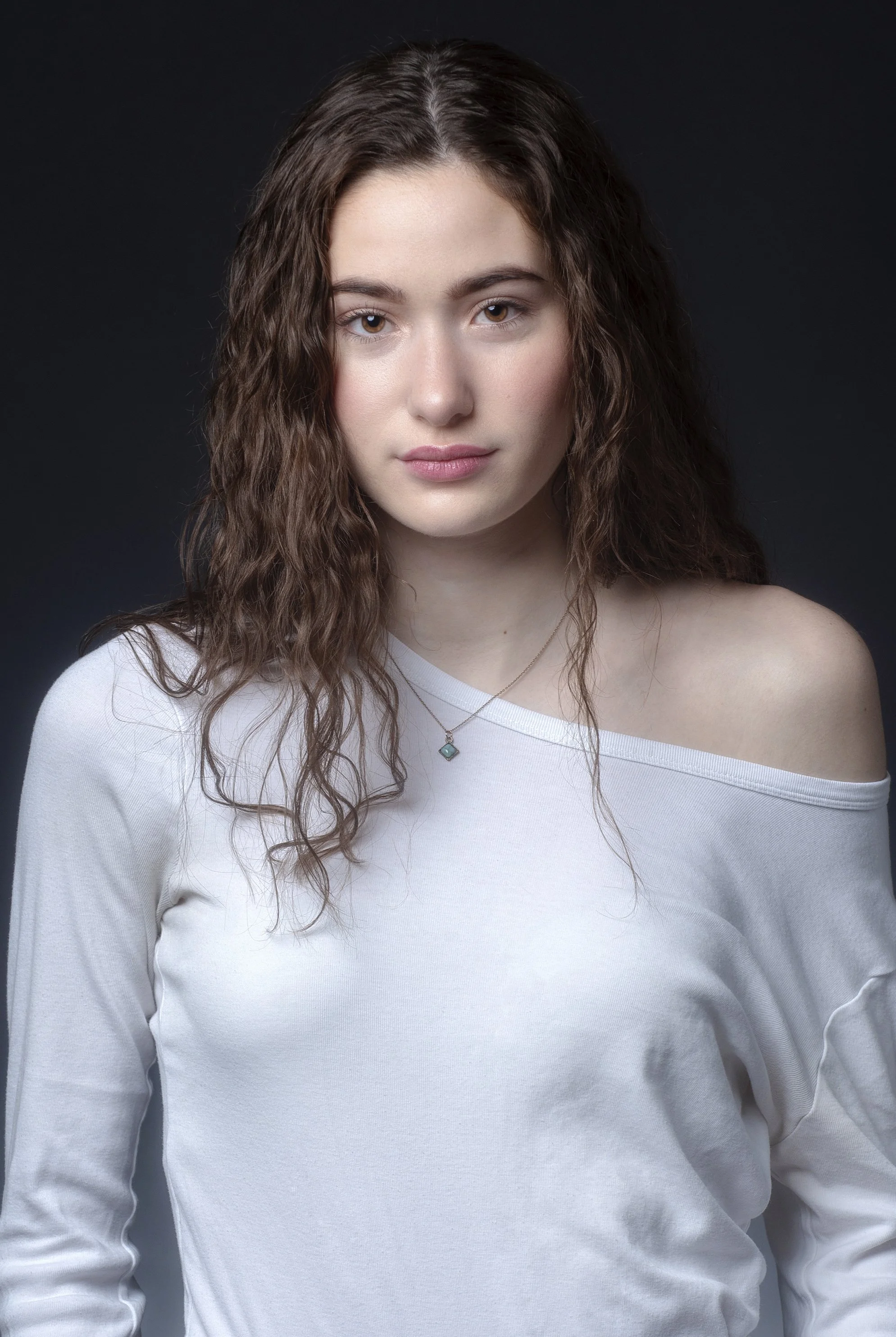 A young woman with curly brown hair wearing a white off-shoulder top and a small necklace, looking directly at the camera against a dark background.