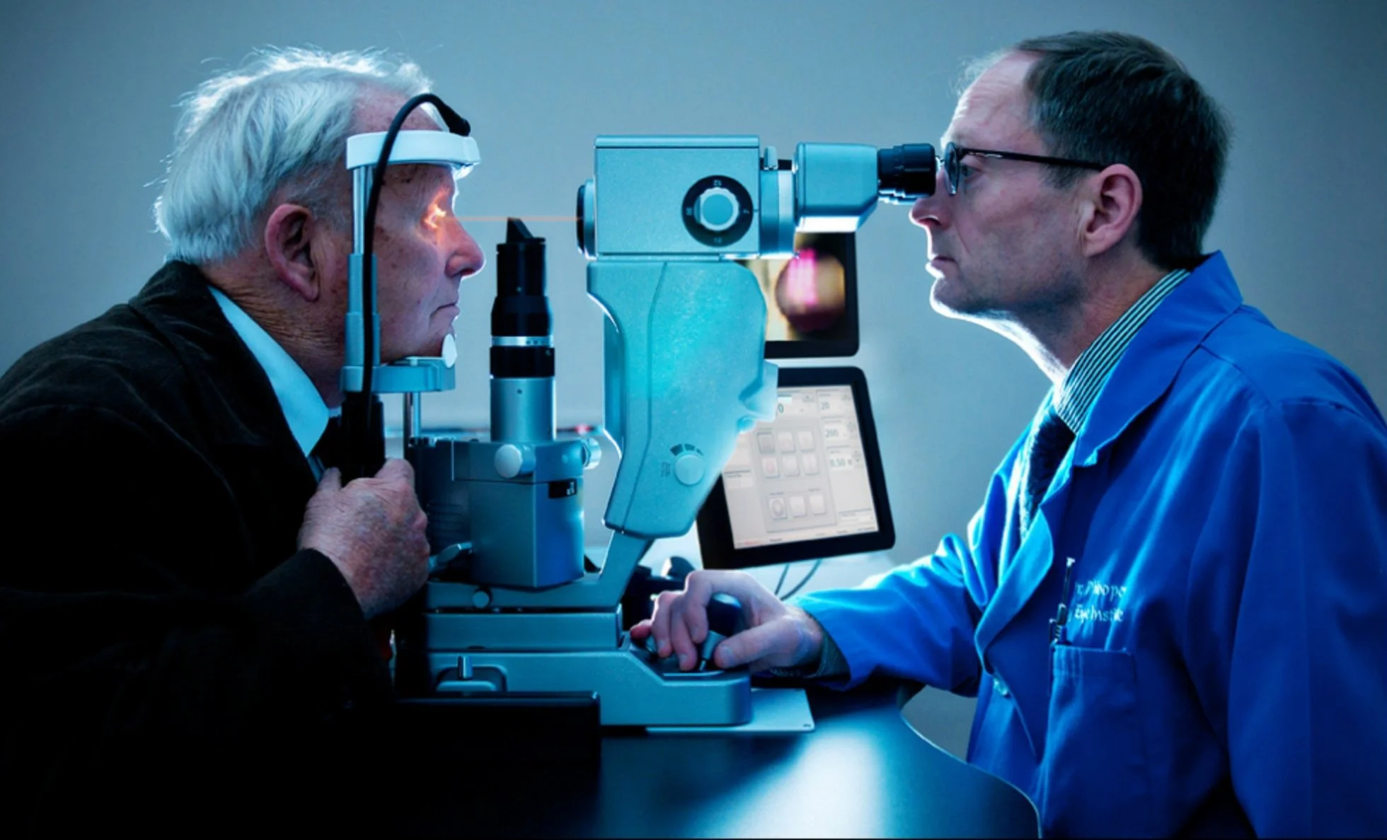 A doctor and an elderly man face an eye examination machine in a clinical setting, with both focusing on the machine's eyepiece.