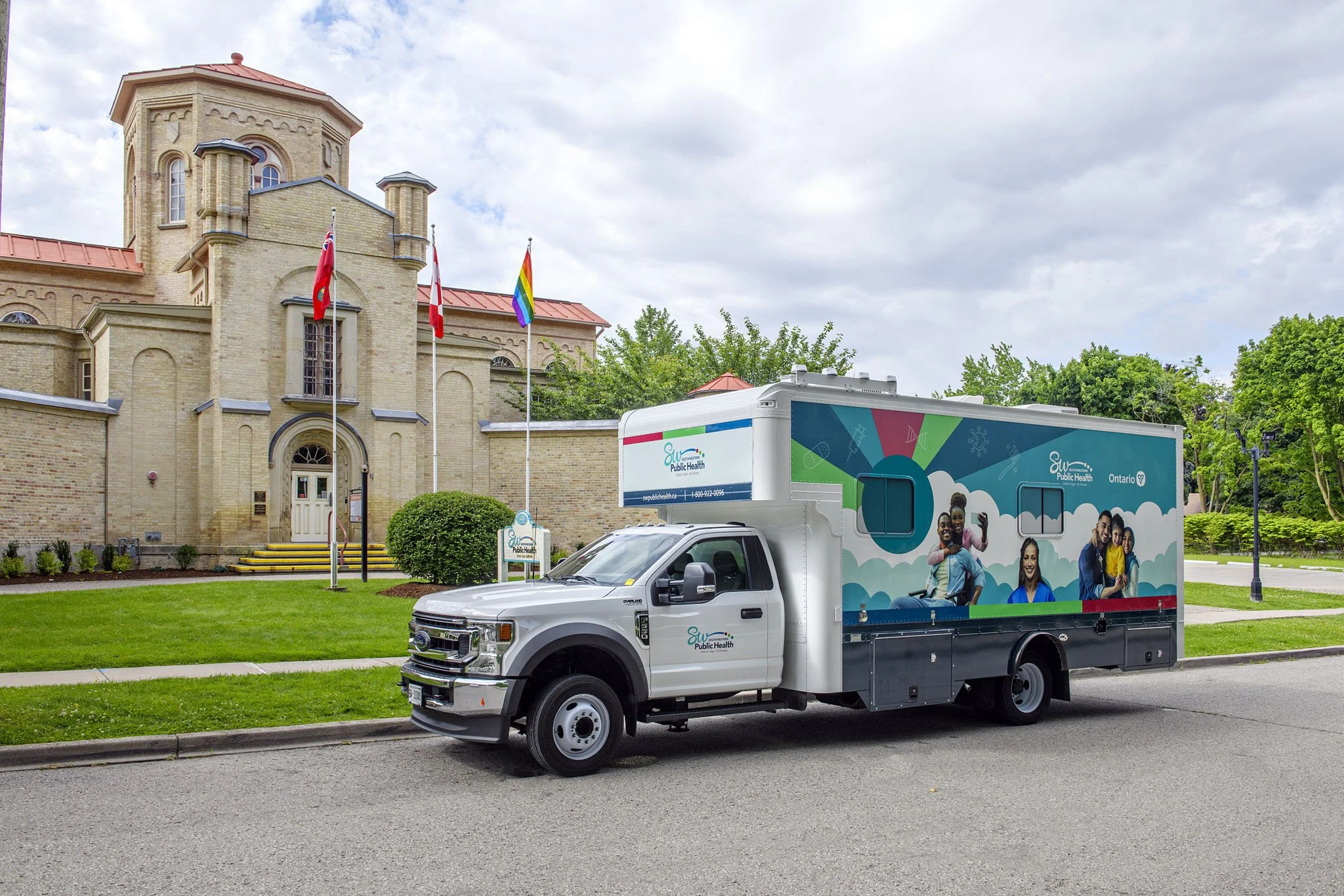 A white truck with colorful graphics and images of diverse people on it, parked on a city street in front of a large brick building with flags, trees, and a blue sky with clouds.