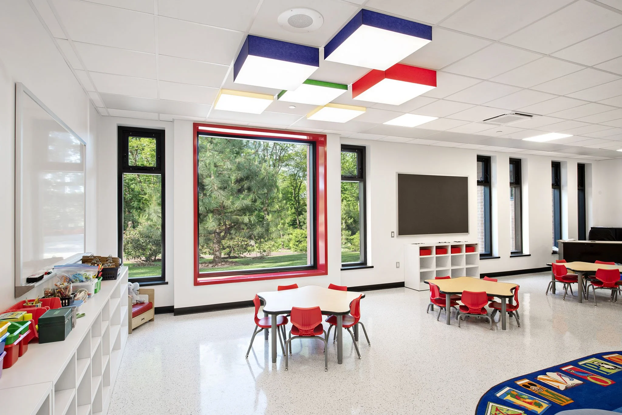 A modern classroom with large windows showing a lush green outdoor scene. The ceiling has colorful square light fixtures. There are small red chairs around multiple round tables and a white bookshelf with various supplies. The room appears bright and