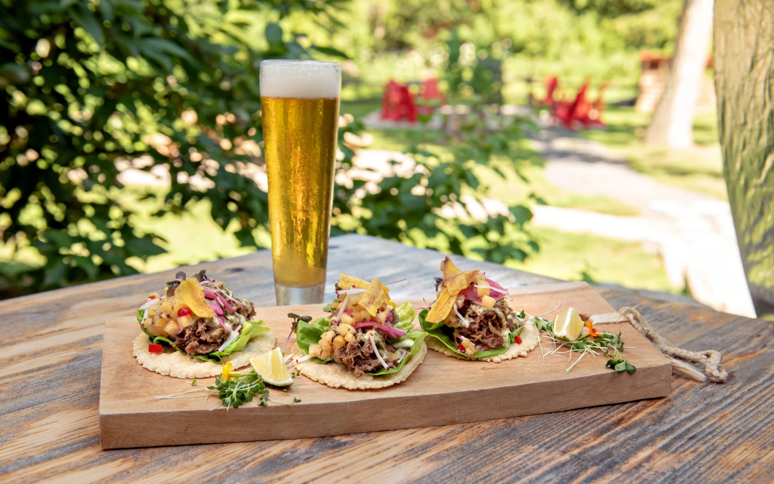 Tacos with shredded meat, vegetables, and lime wedges on a wooden serving board outdoors, with a glass of beer in the background.