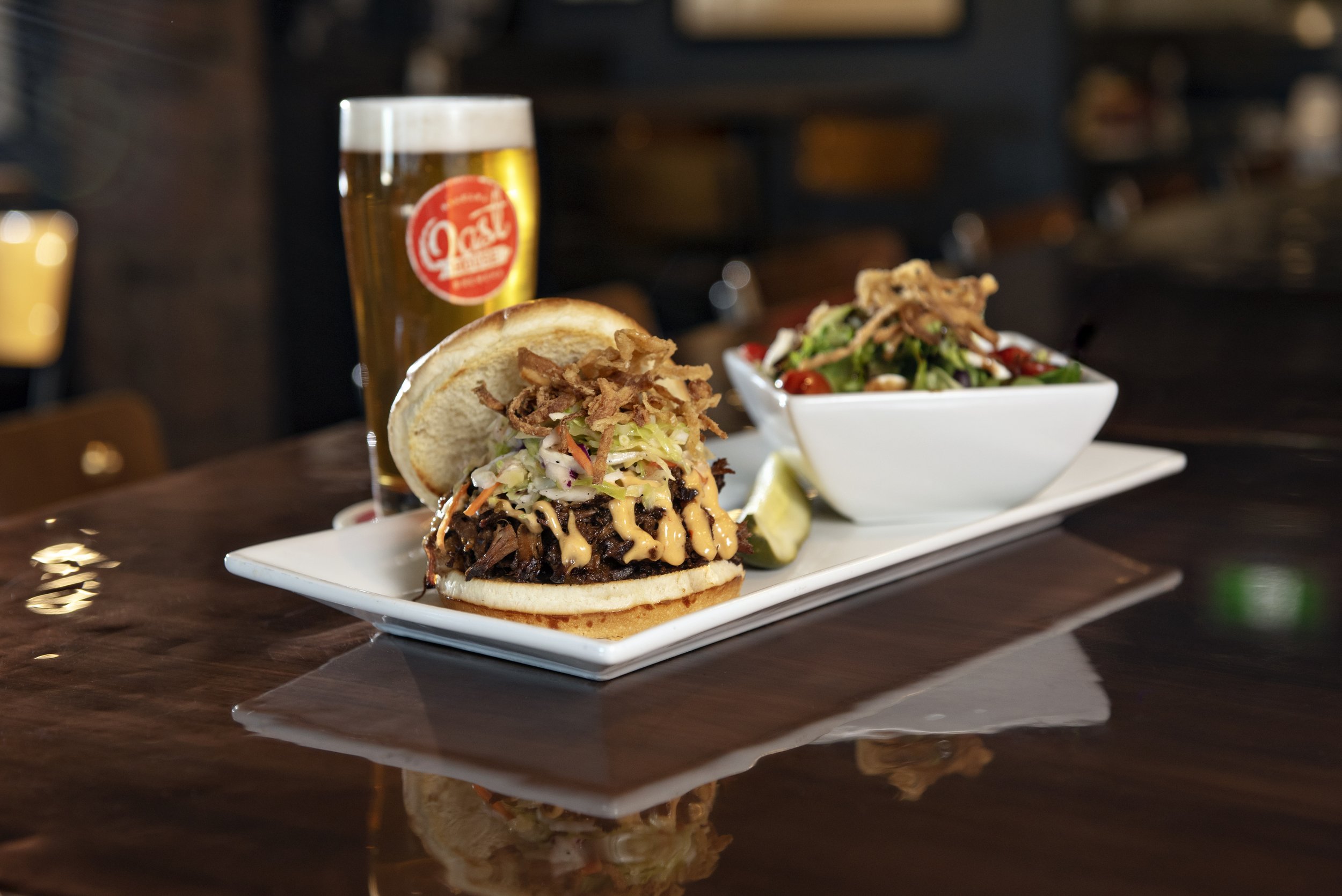 A plate with a sandwich filled with shredded meat, coleslaw, and fried onions, accompanied by lemon wedges. In the background, there is a bowl of salad with greens and cherry tomatoes, and a glass of beer with a foamy head and a red and white logo.