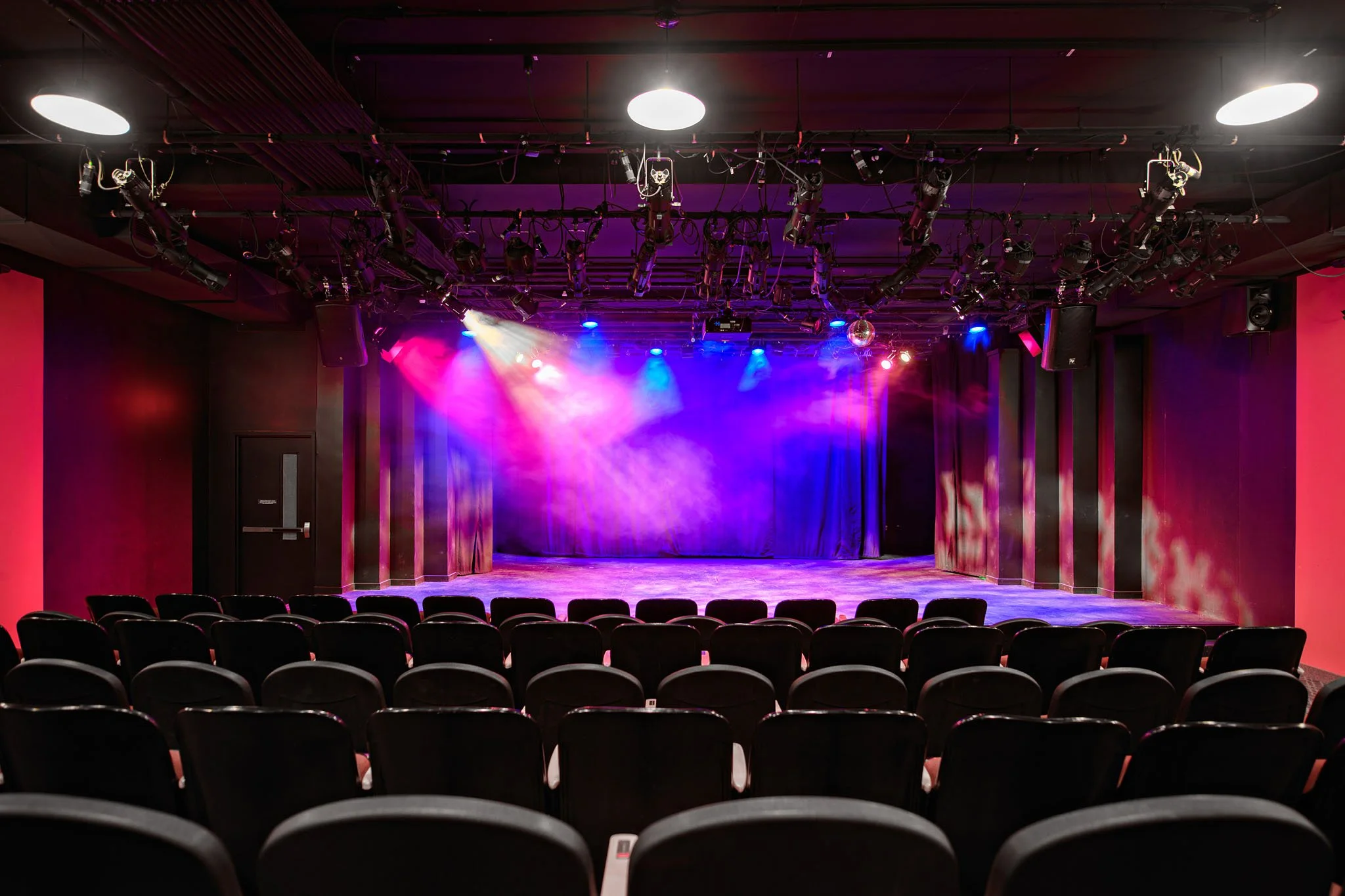Empty small theater with rows of black chairs and a stage illuminated with colorful lights.