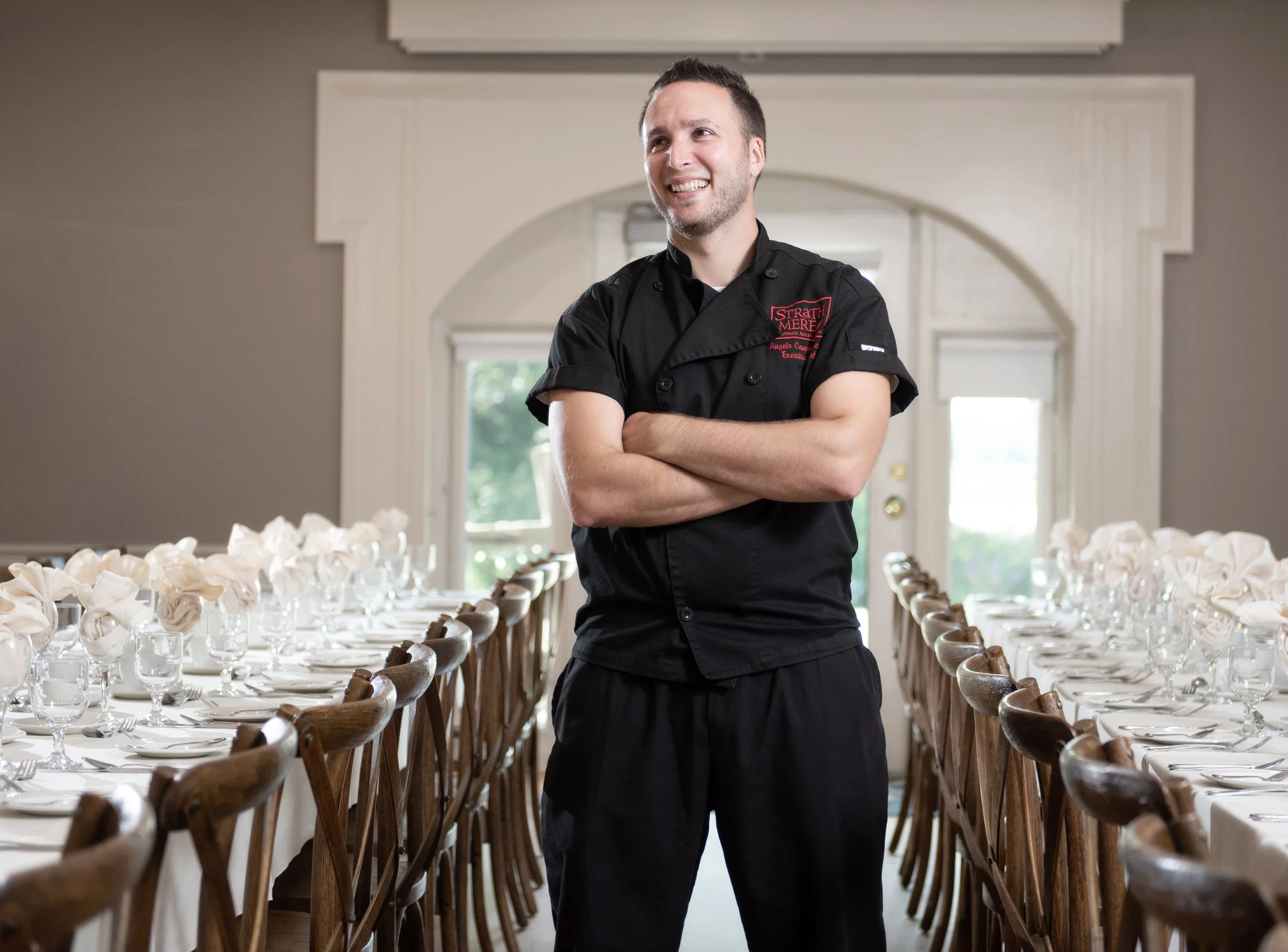 A smiling man in a black chef's jacket standing with arms crossed in a dining room with a long set table and wooden chairs
