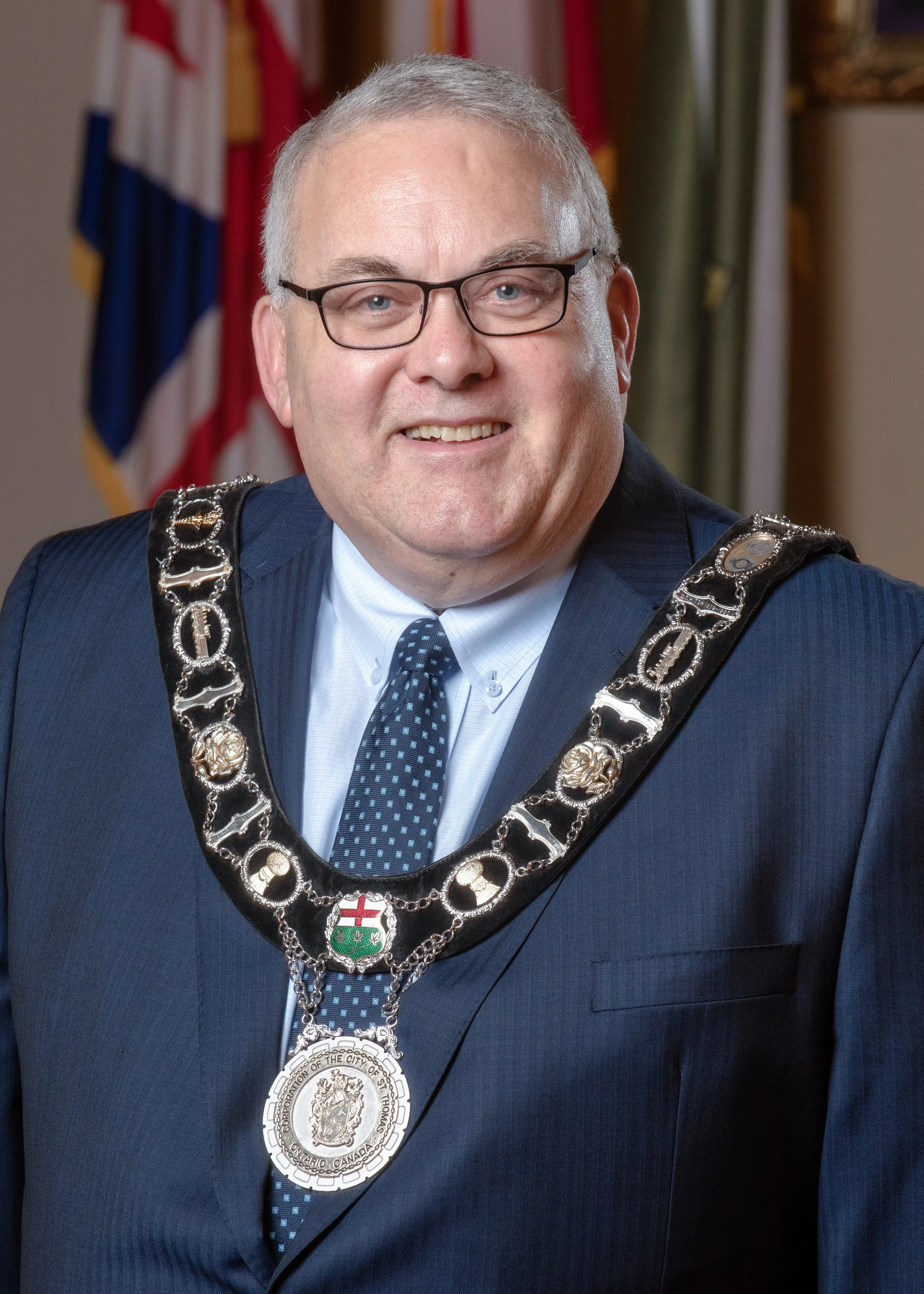 A man in a suit and tie wearing a city mayor's chain with a large medallion, standing in front of flags.