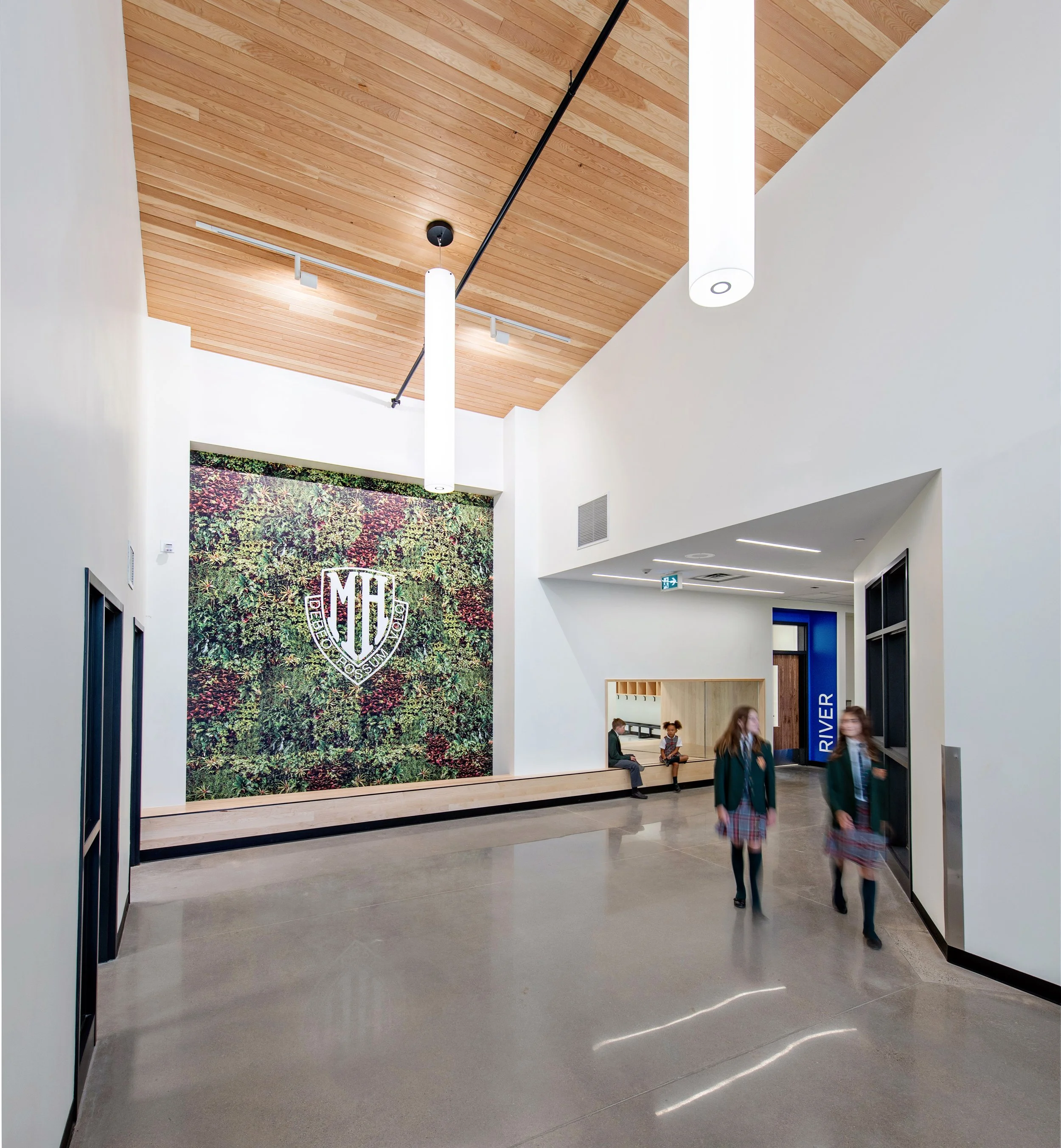 Modern school hallway with a wood-paneled ceiling, large wall decor featuring a plant pattern and a crest, and students walking and sitting around.
