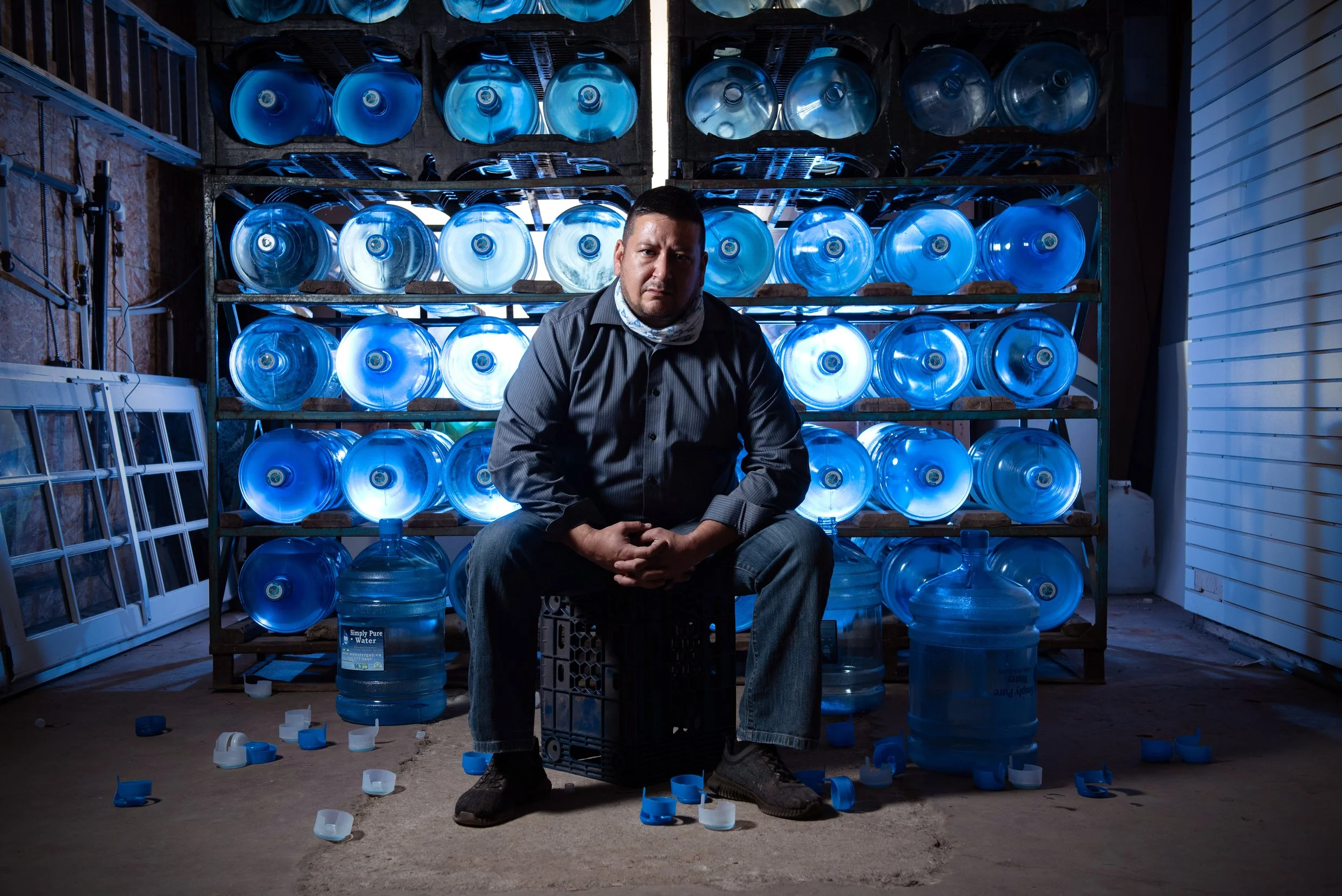 A man sitting on a milk crate in front of shelves filled with large water bottles illuminated with blue light, with scattered bottle caps on the floor in a storage or warehouse setting.