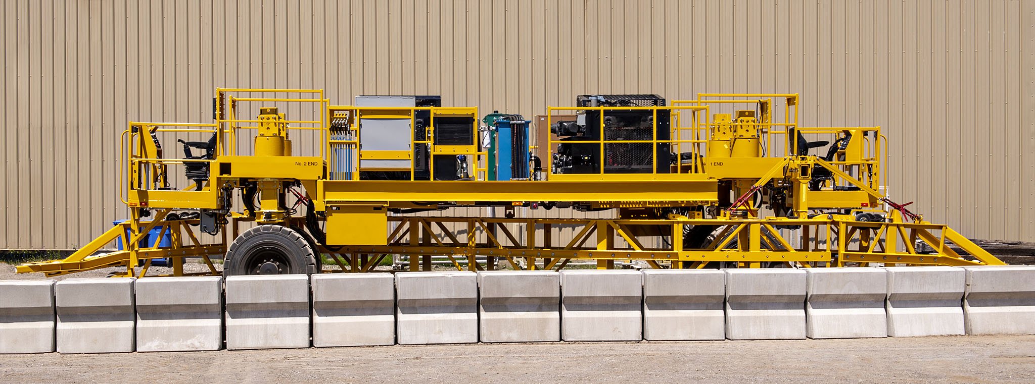 Yellow construction or industrial vehicle parked outdoors with concrete barriers in front, against a beige metal building background.