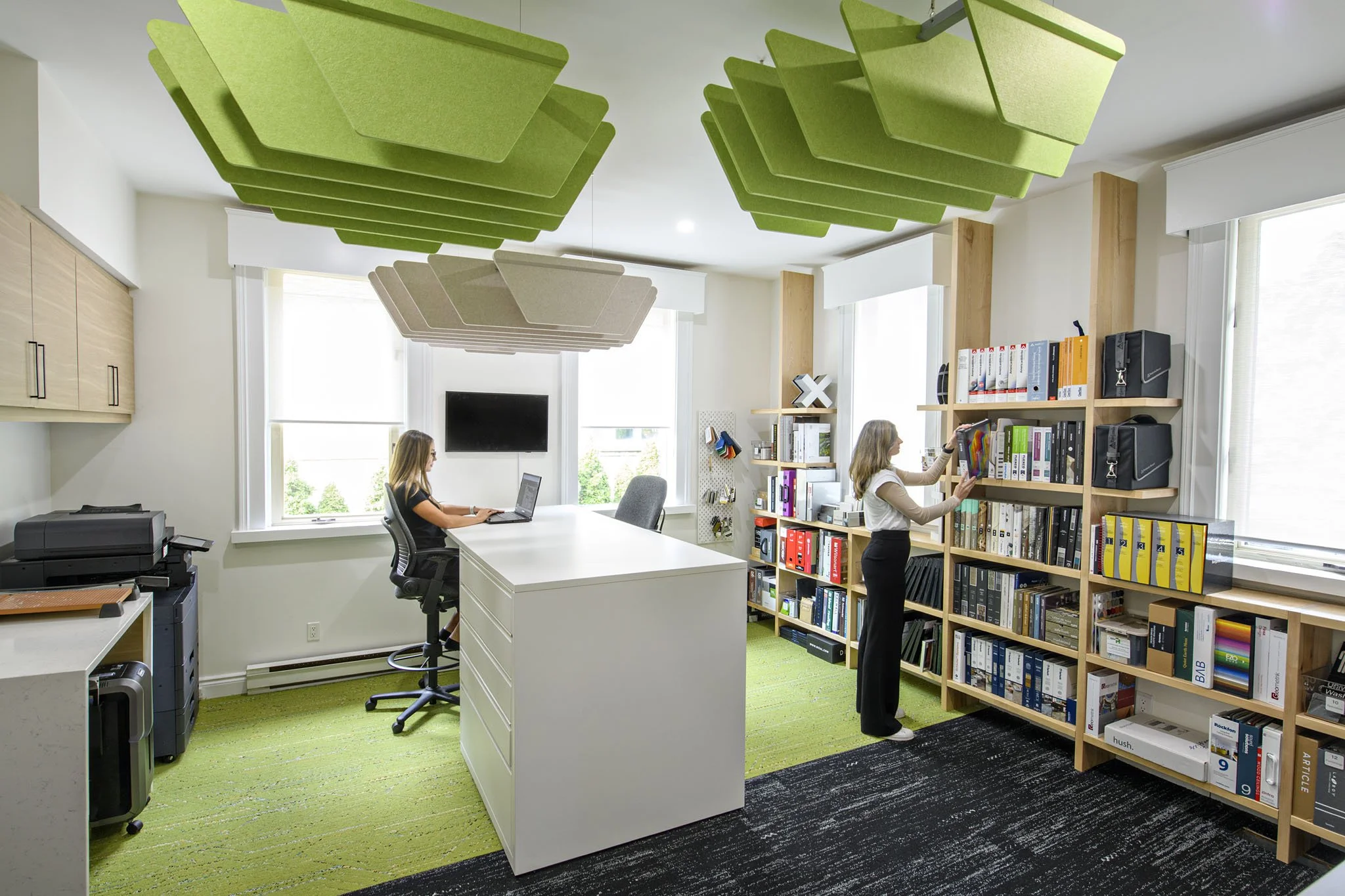 Office with two women, one working at a desk on a laptop and the other browsing books on a bookshelf, bright space with large windows, green and black carpet, unique ceiling acoustic panels in green and beige.