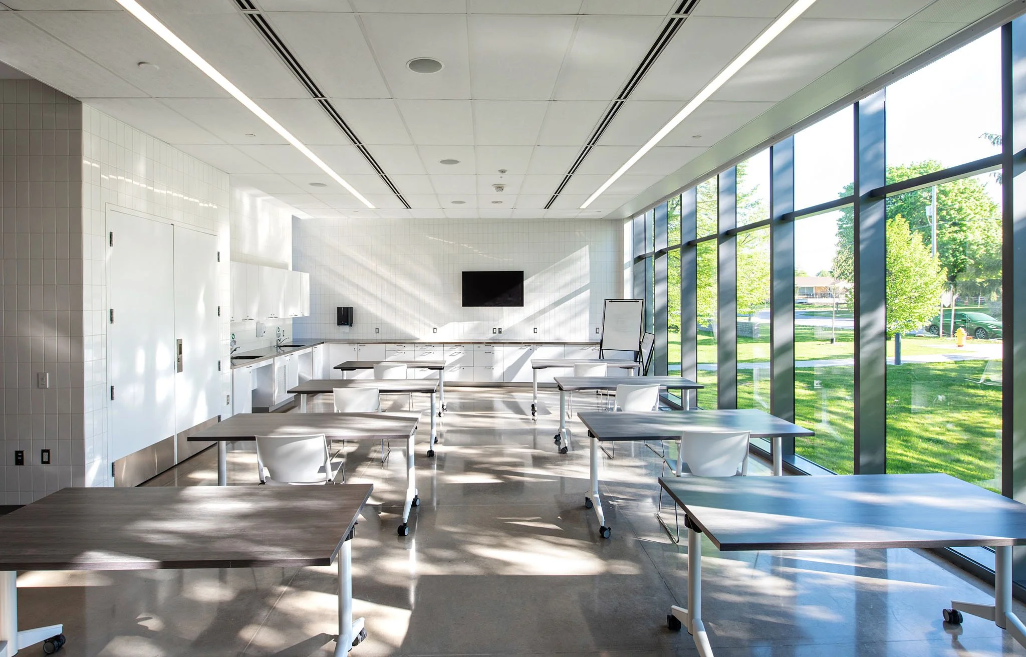 A modern conference room with white chairs and tables, large glass windows showing trees and a parking lot outside, and a wall-mounted television.