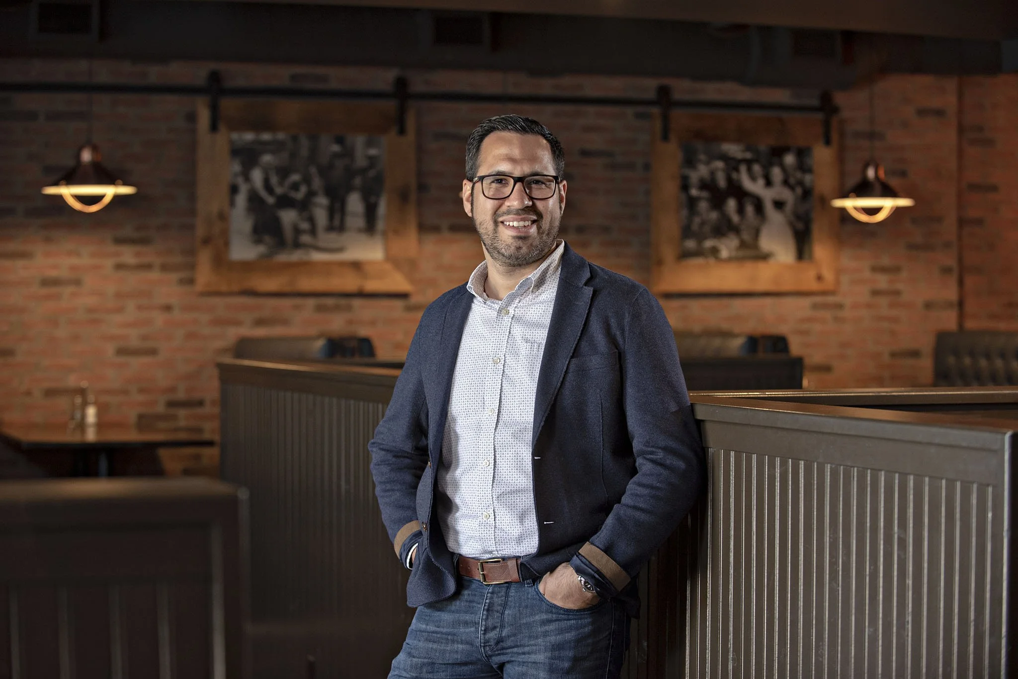 A man in glasses, dressed in a blazer and dress shirt, smiling and standing in a restaurant with exposed brick walls, framed black and white photos, and warm lighting.