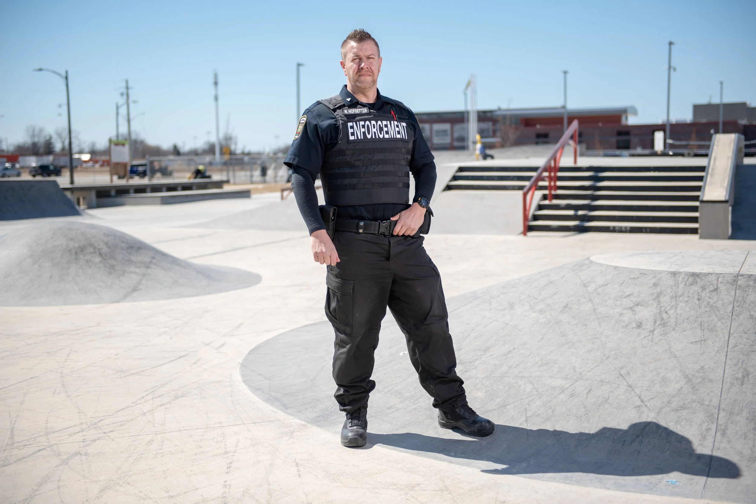 A police officer wearing a black vest with 'ENFORCEMENT' written on it stands at a skate park on a sunny day. The background shows ramps and stairs at the skate park and a building, with a clear blue sky overhead.