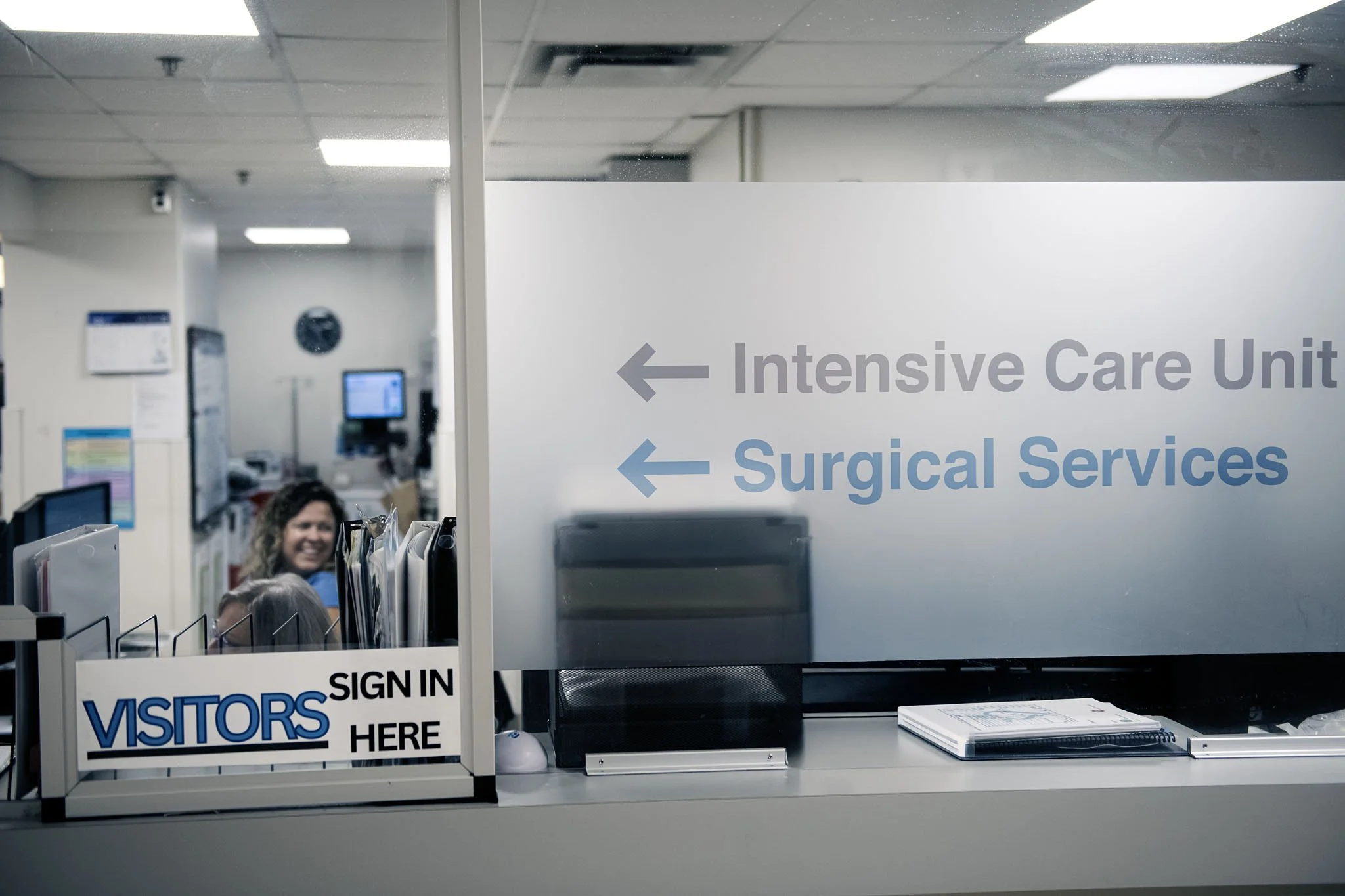 View of a hospital reception or registration area with signs indicating directions to Intensive Care Unit and Surgical Services, a sign instructing visitors to sign in here, and a smiling woman behind a counter.