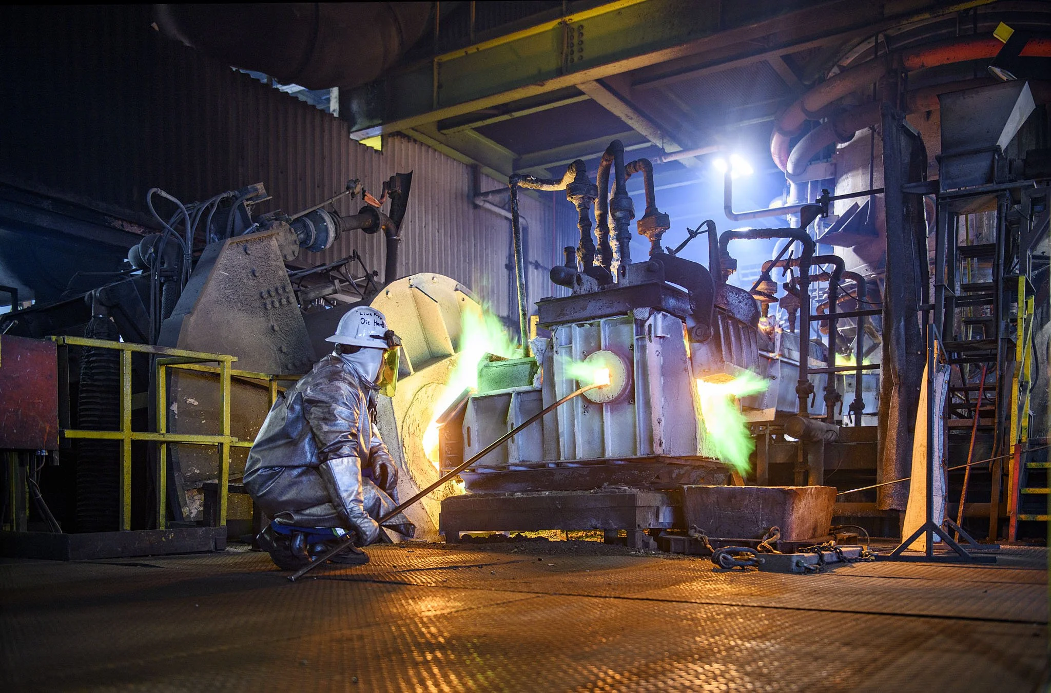 A worker in protective clothing and helmet working in a steel foundry with molten metal pouring from a furnace, surrounded by industrial pipes and equipment.