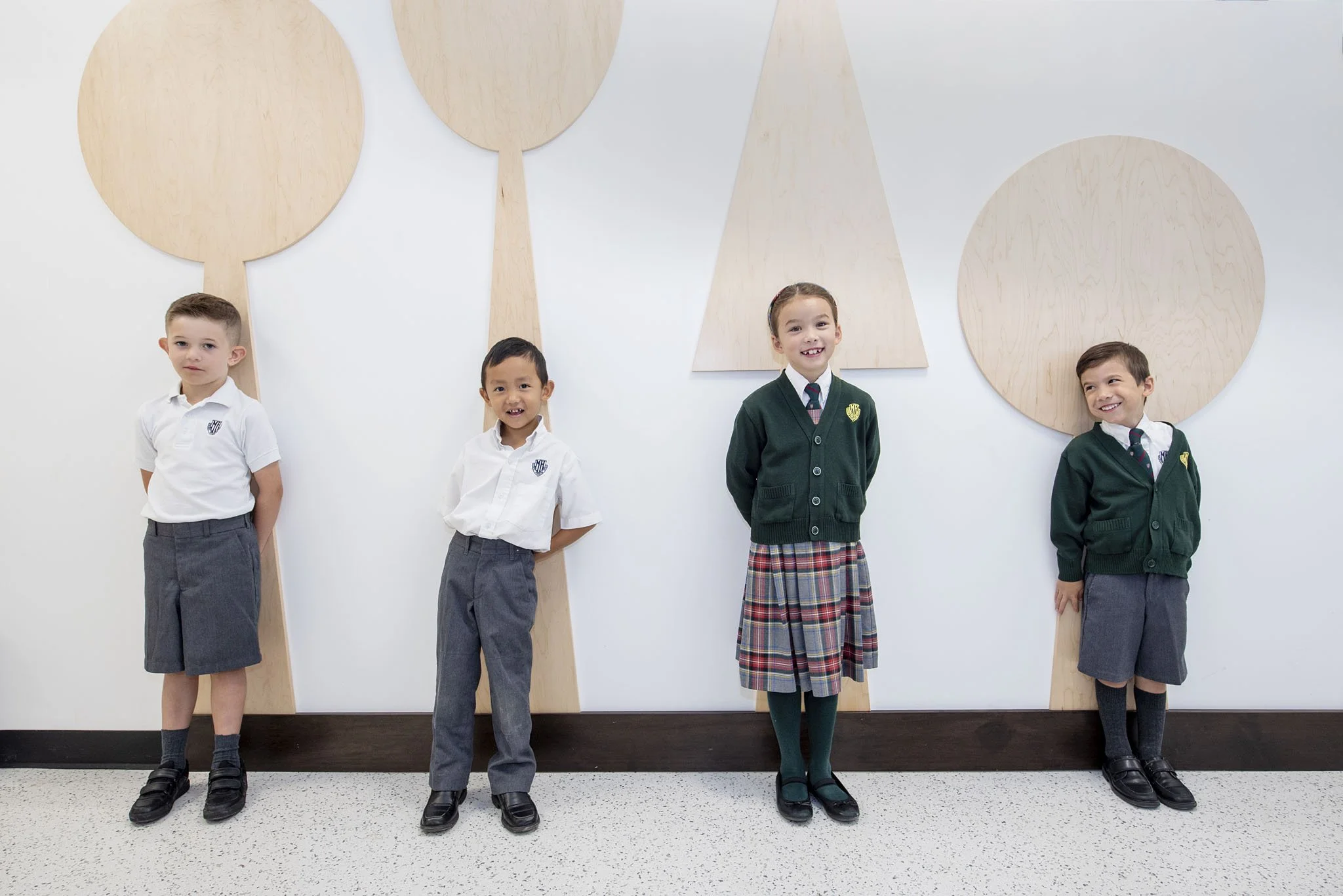 Four young children in school uniforms standing side by side against a white wall with large wooden abstract art pieces. The children are smiling and looking at the camera.