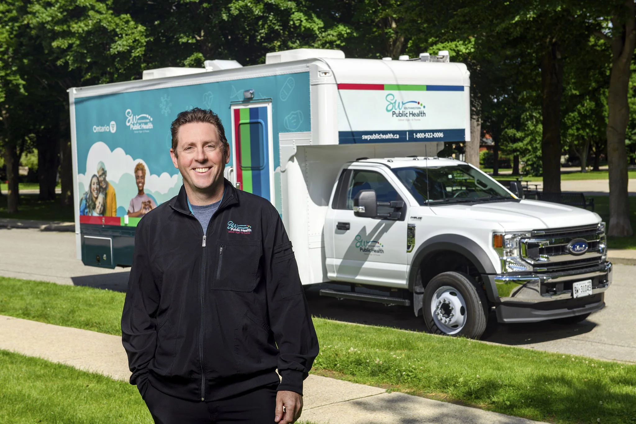 A smiling man in a black jacket standing outdoors in front of a mobile health unit trailer and a truck, surrounded by green trees and grass.
