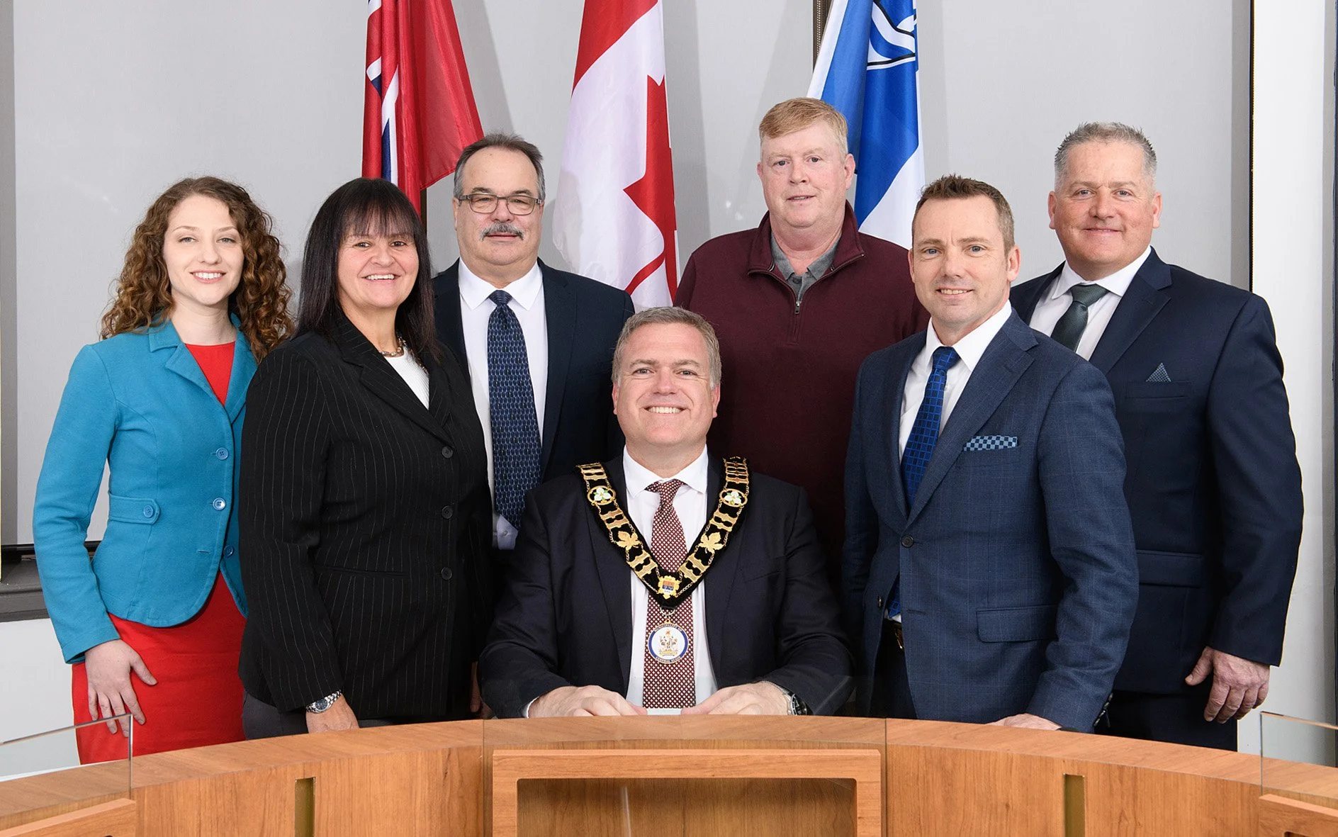 A group of eight individuals, including a man in a suit with ceremonial chains, posing for a photo indoors with flags of Canada, the United Kingdom, and other countries in the background.