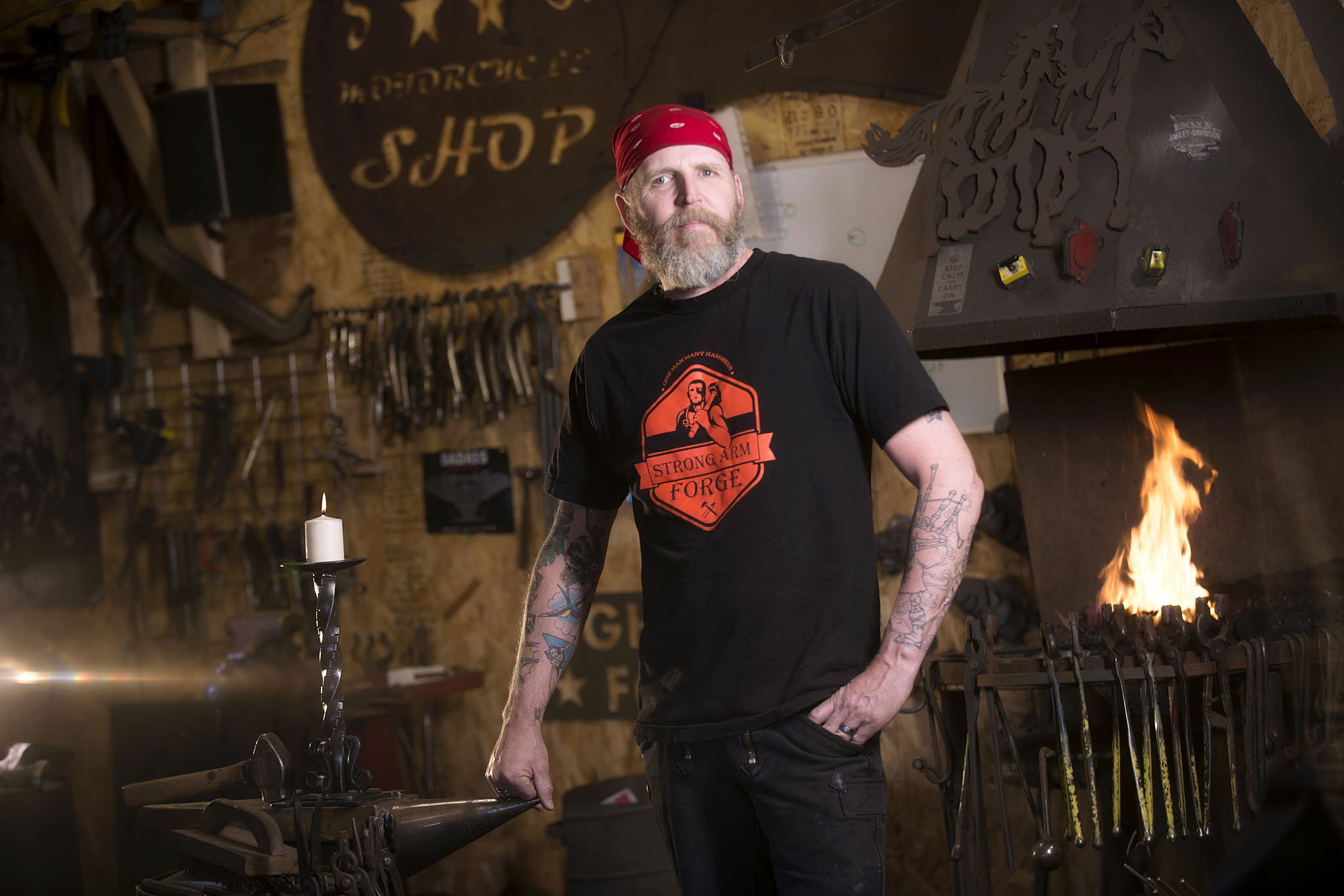 A man with a beard, tattoos, wearing a red bandana and a black T-shirt with a logo, stands in a blacksmith workshop. Behind him are tools. There is a fire burning in the forge on the right side of the image. Executive portrait photography. 