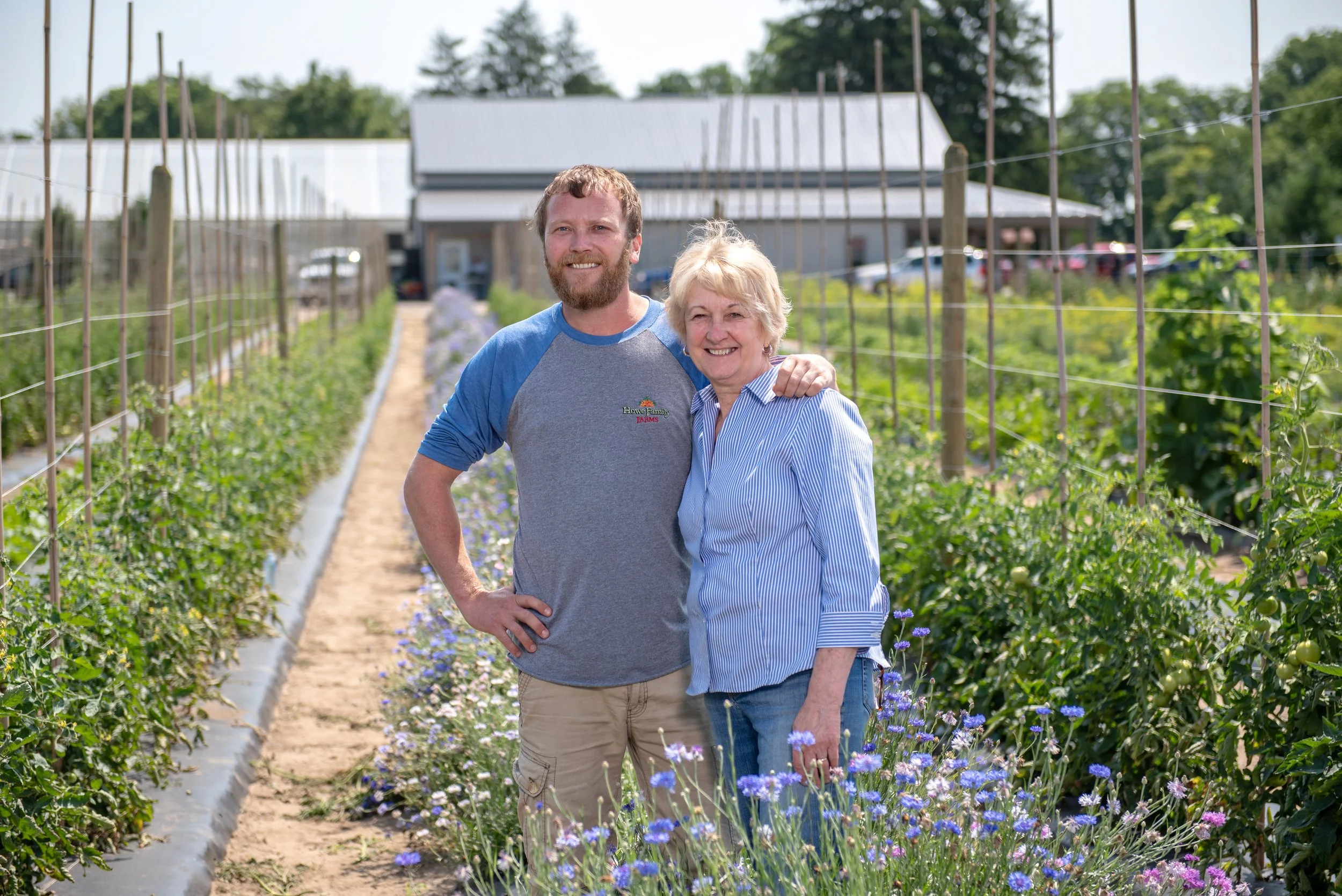 A man and an older woman standing together in a garden with rows of flowering plants and a greenhouse building in the background, on a sunny day.