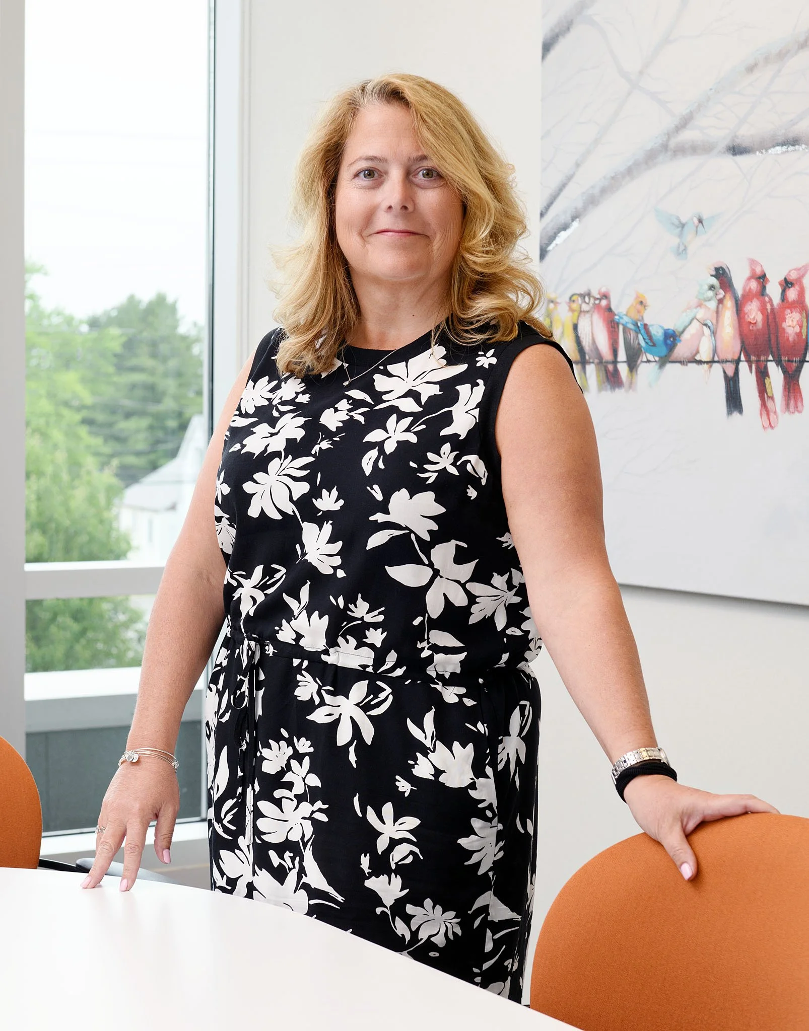 Woman with blonde hair wearing a black dress with white floral pattern, standing in a bright room with large windows and a colorful bird art piece on the wall, resting her hand on an orange chair.