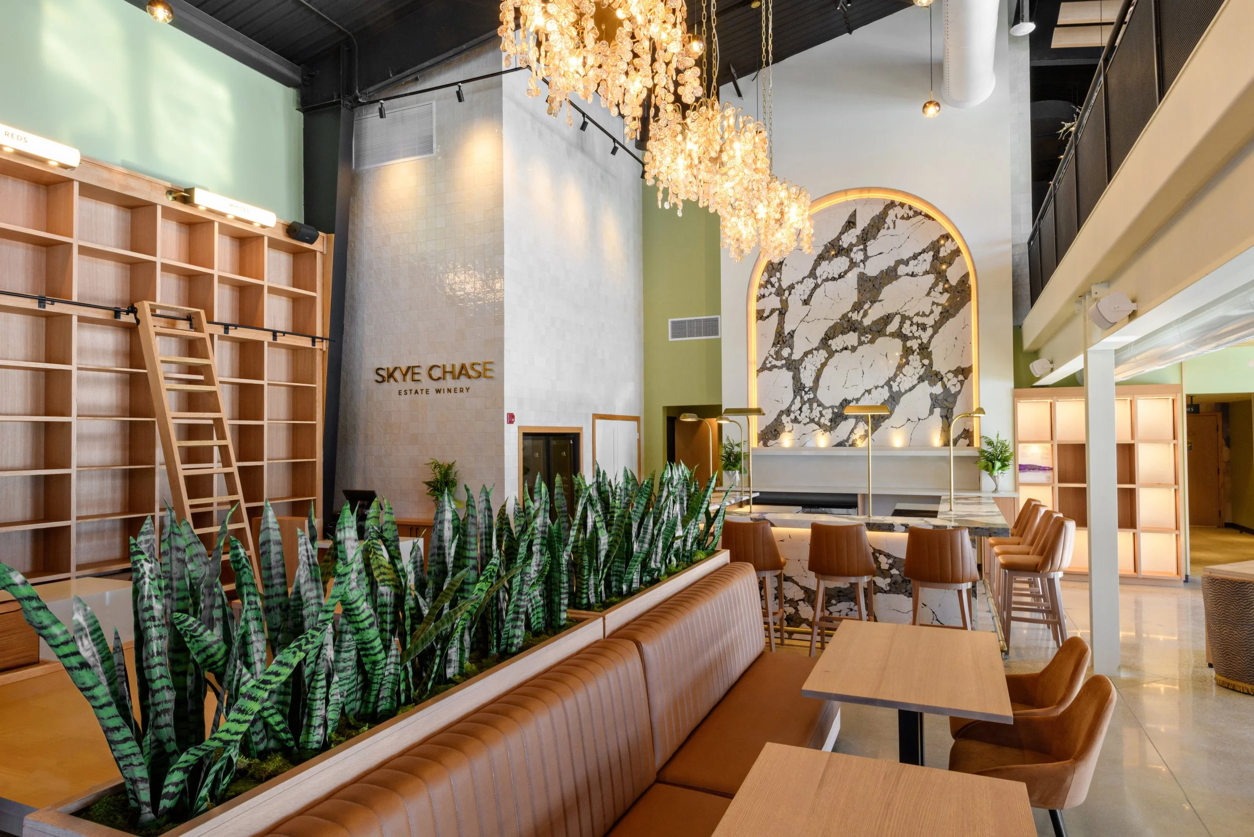 Modern interior of a winery lobby with a high ceiling, a large marble wall with backlighting, and an elaborate chandelier made of glass or crystal. There are wooden shelves and a lounge area with tan leather seating and wooden tables. Green plants li