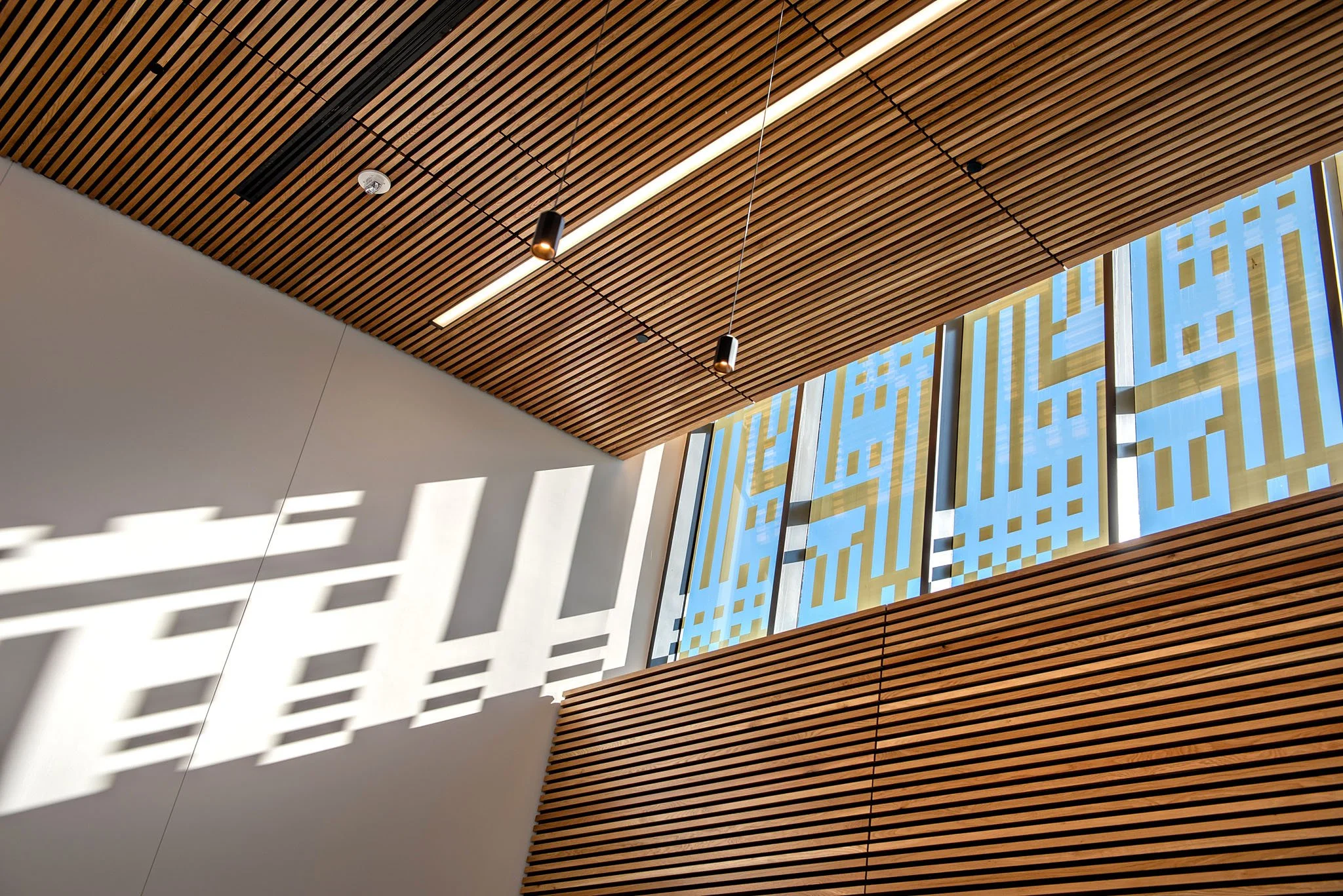 Interior view of a modern building ceiling with wooden slats, pendant lighting, and large windows with decorative yellow and blue patterns.