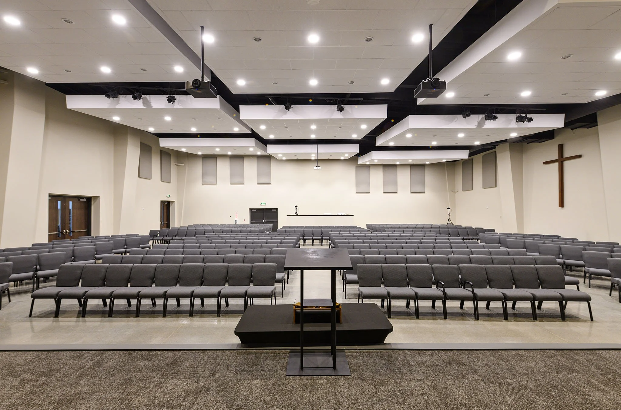 Empty church sanctuary with gray chairs arranged in rows facing a stage with black steps, a cross on the wall, and ceiling lights.