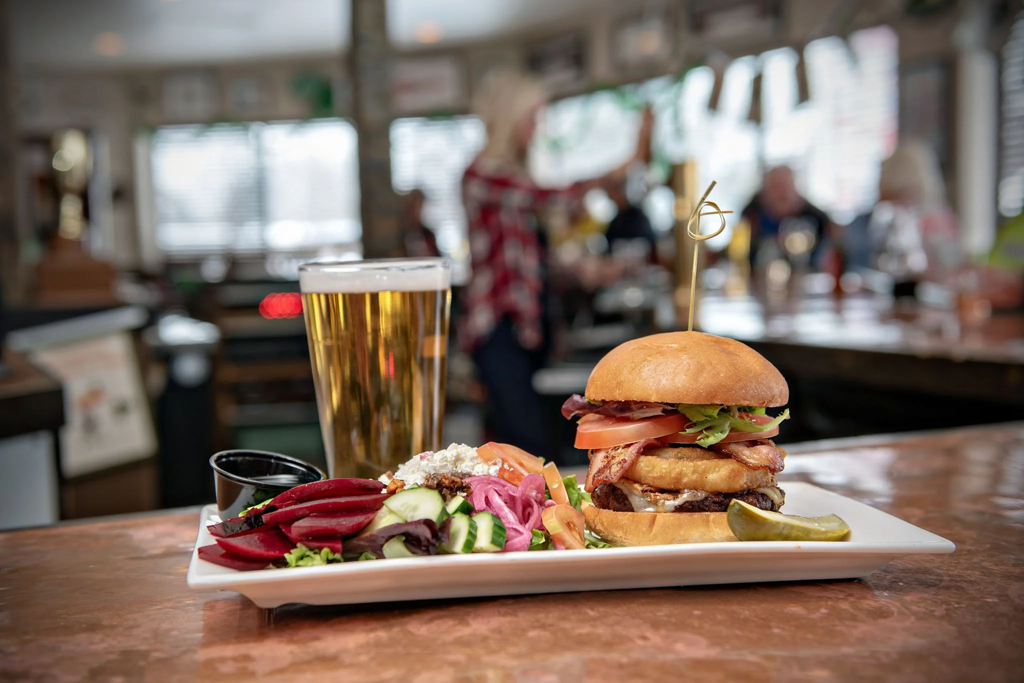 A plate with a burger, salad, and pickles, a tall glass of beer, and a small cup of sauce on a wooden table in a restaurant with large windows and blurred customers in the background.