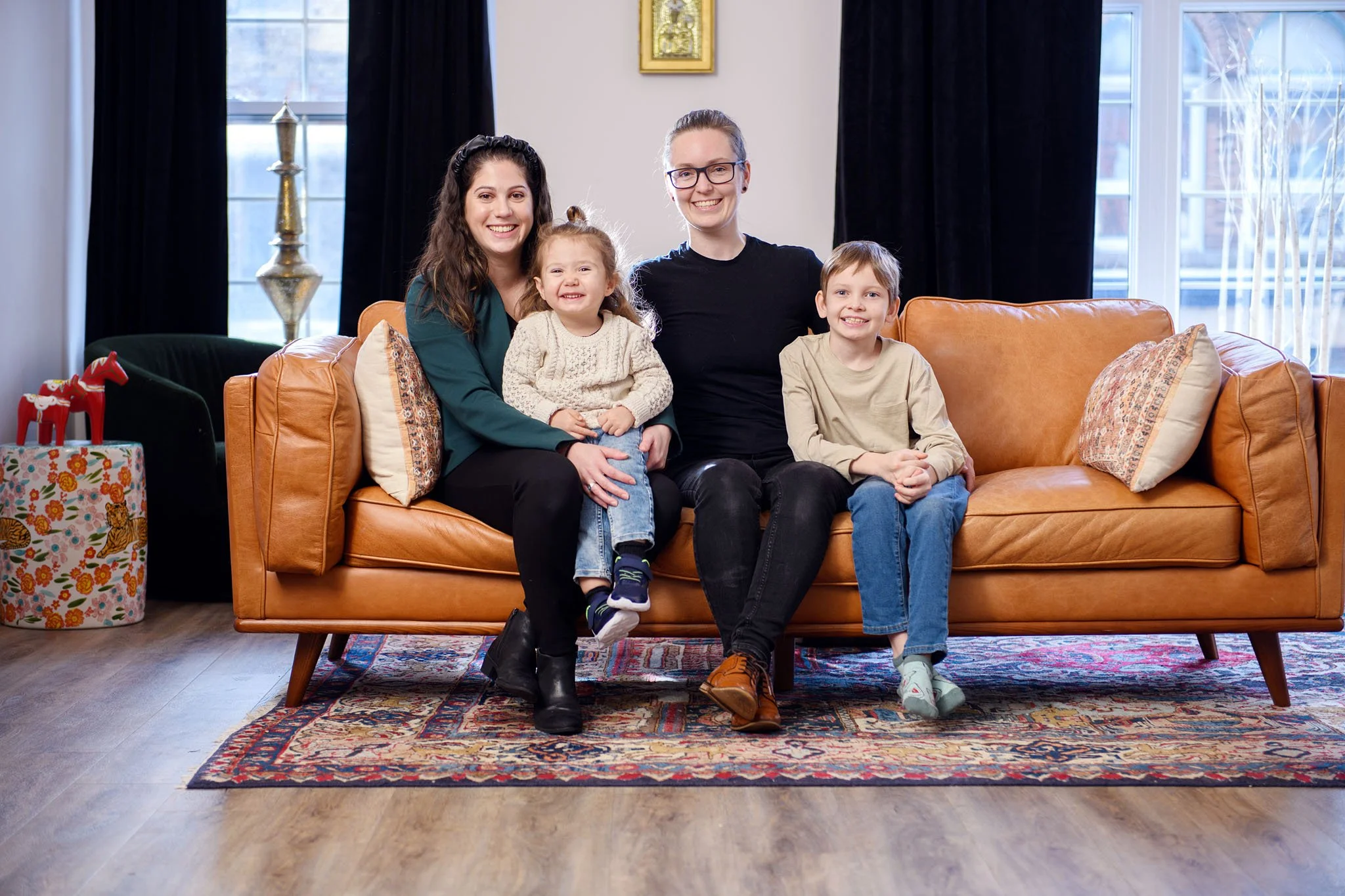 A family of four sitting on a tan leather couch in a living room, smiling at the camera. The family includes two women, a young girl, and a young boy. Behind them are large windows with black curtains, and on the left side, there's a gift box with a 