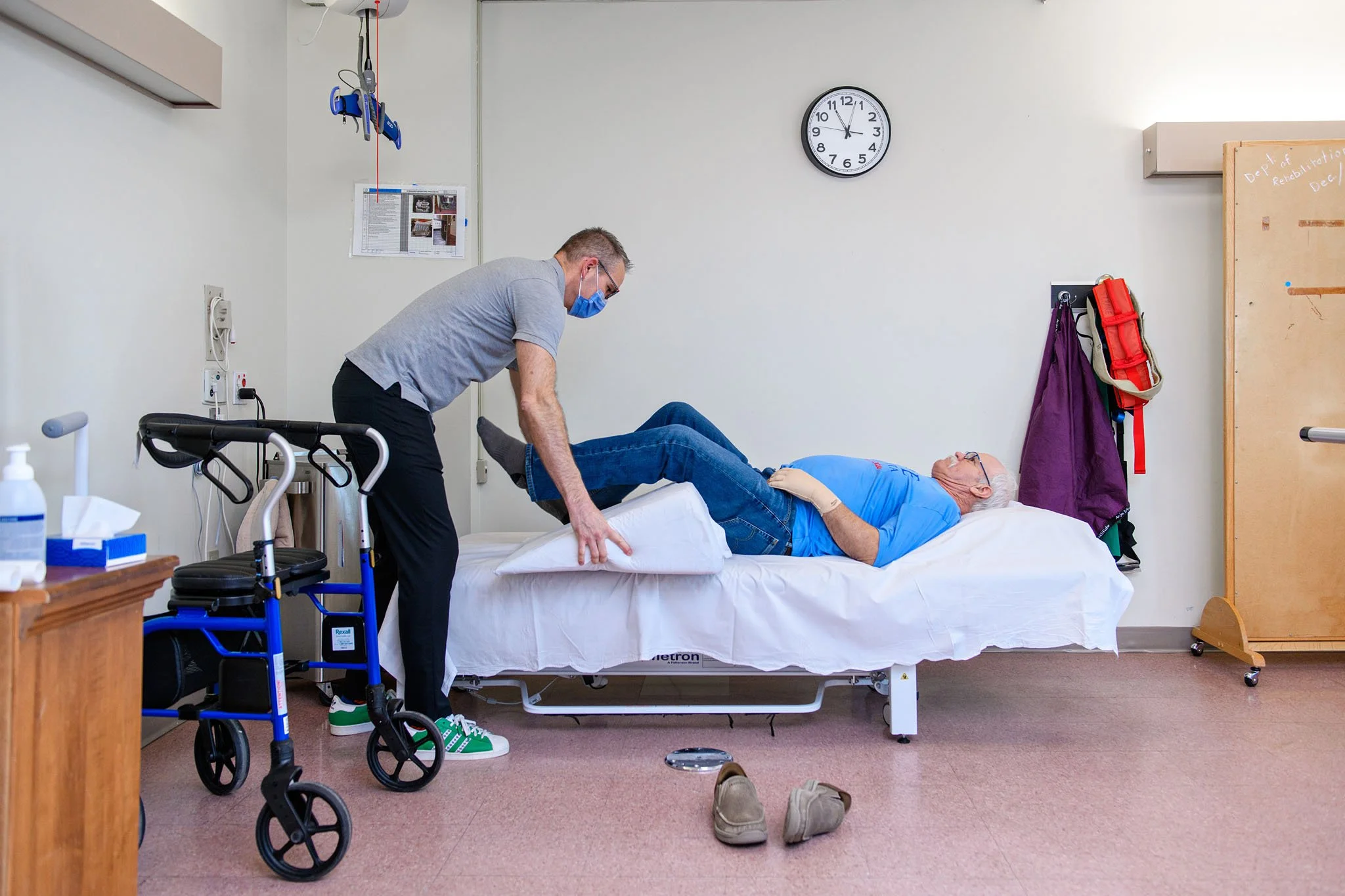 A healthcare room with a patient lying on a hospital bed receiving care from a nurse. The patient is elderly, wearing glasses and a blue shirt. The nurse is wearing a gray shirt, glasses, and a face mask. Medical equipment such as a walker, gloves, a