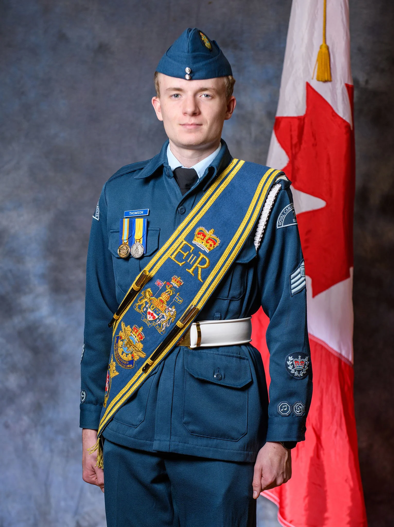 A young man in a blue military uniform with medals and a sash, standing in front of a Canadian flag.