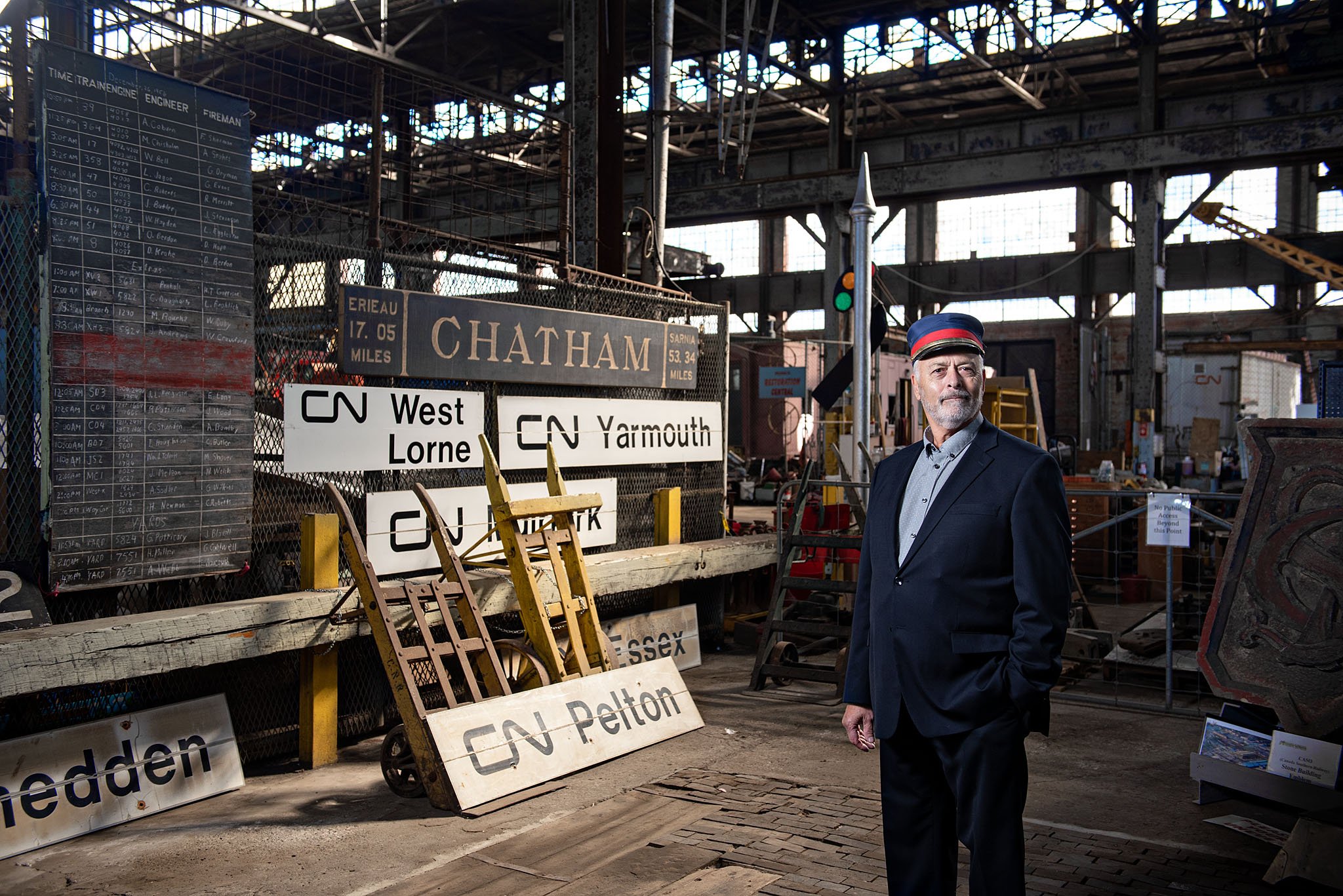 A man in a suit and cap standing inside a historic train station with signs for various routes and destinations, including Chatham and Salem, and a timetable board on the left. Executive portrait photography