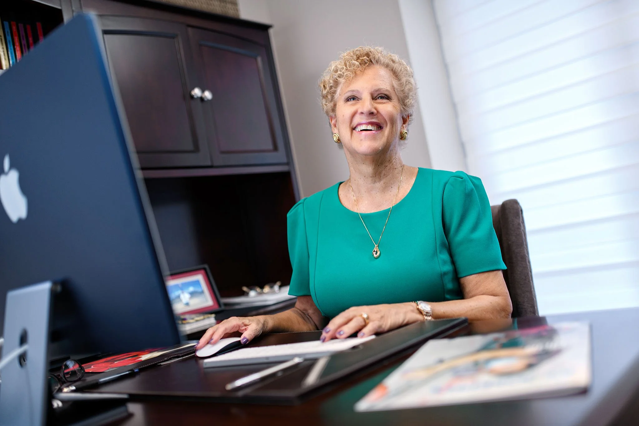 A cheerful older woman with curly blonde hair, wearing a teal dress and gold jewelry, working on a computer in a bright office.