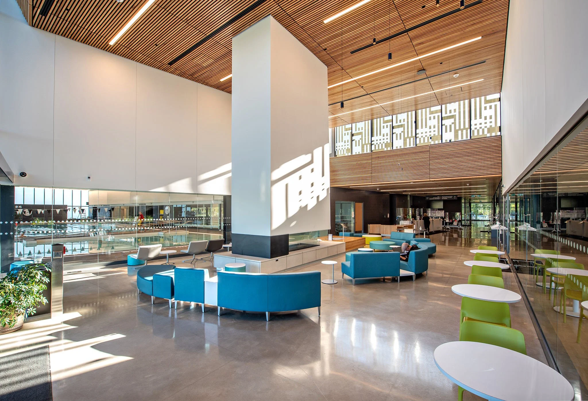 Modern indoor space with high wooden ceilings, white walls, and large windows allowing sunlight. There are seating areas with blue and green chairs, small white tables, and a few people. In the background, there is a swimming pool visible through a g