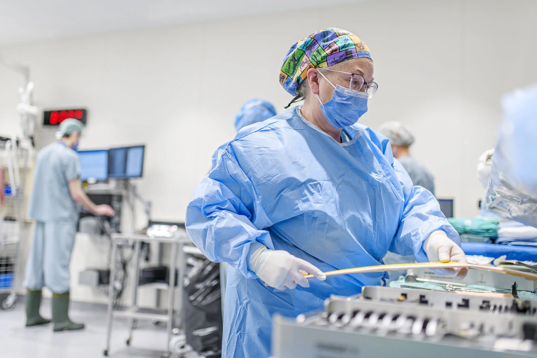 A healthcare worker in blue scrubs, a colorful surgical cap, face mask, and glasses working in a hospital operating room with medical equipment and other staff in the background.