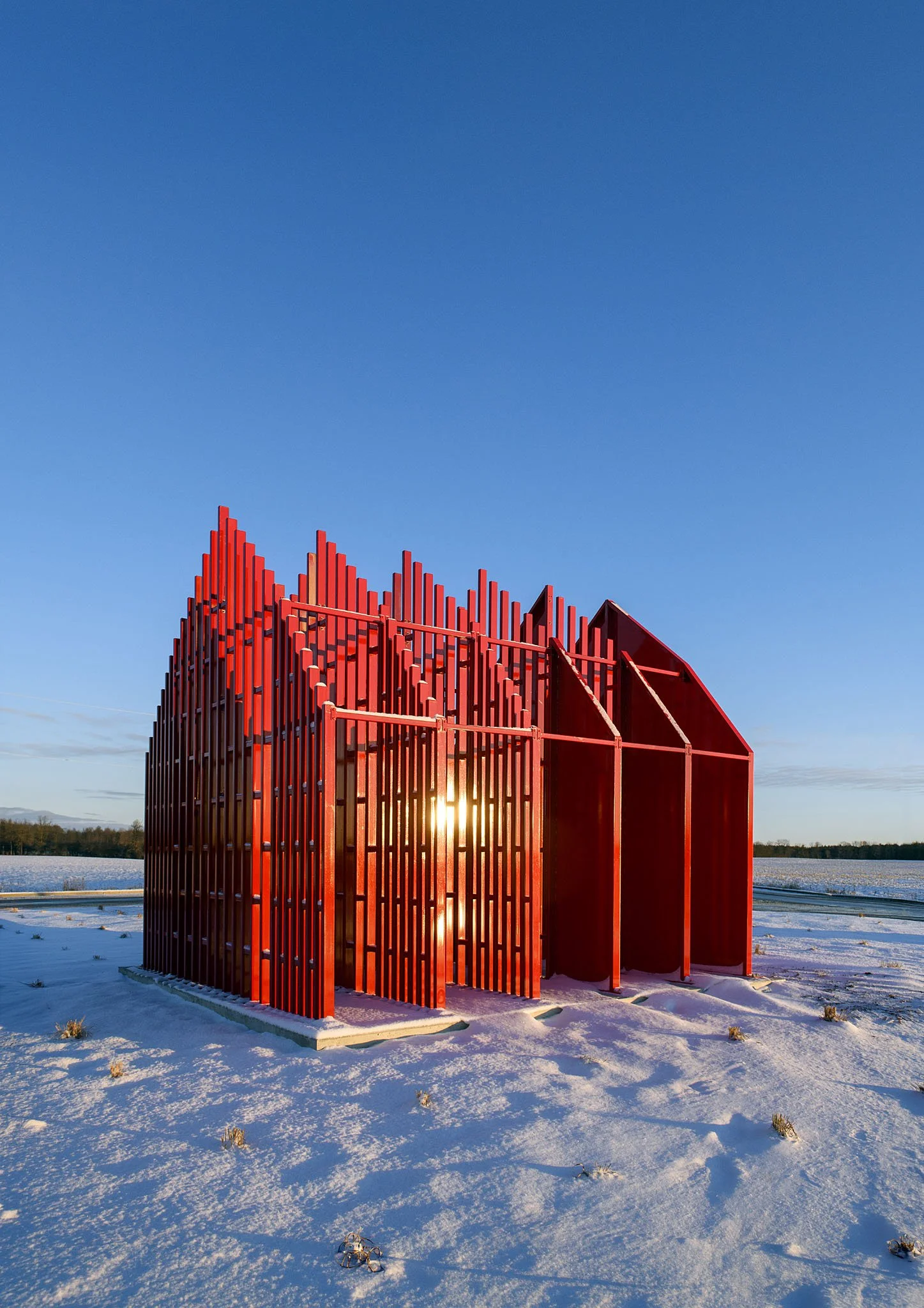 Red modern sculpture with vertical bars in a snow-covered field, with the setting sun behind it and a clear blue sky.