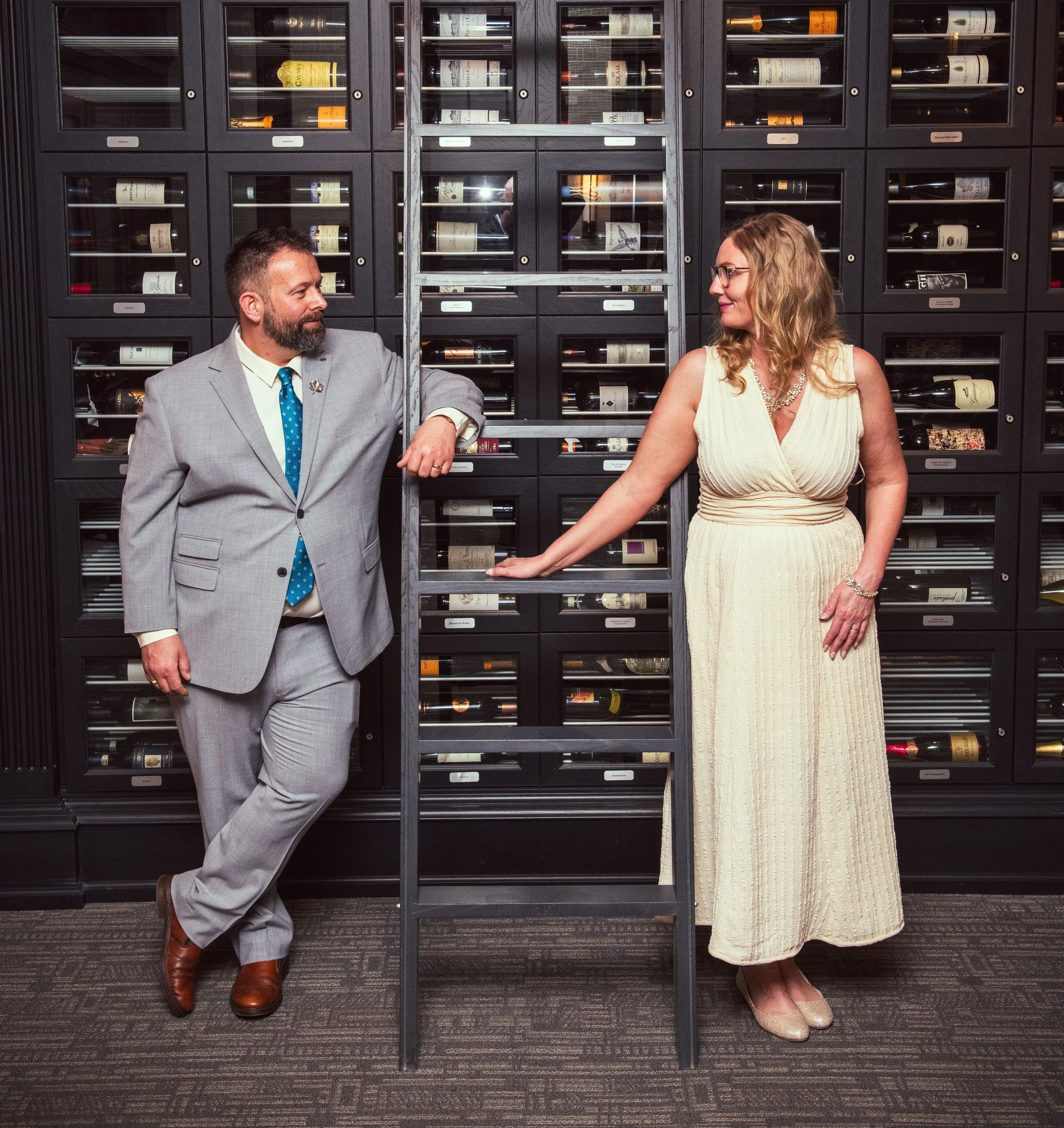 A man in a gray suit with a blue dotted tie and a woman in a beige dress with glasses are standing in front of a wall of wine bottles behind glass doors. They are looking at each other and smiling.
